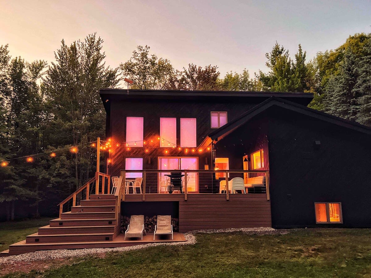 Modern black house with large windows reflecting a pink sunset, featuring a multi-level wooden deck with string lights, outdoor seating, and a grill. Two lounge chairs are visible on the lower deck level.