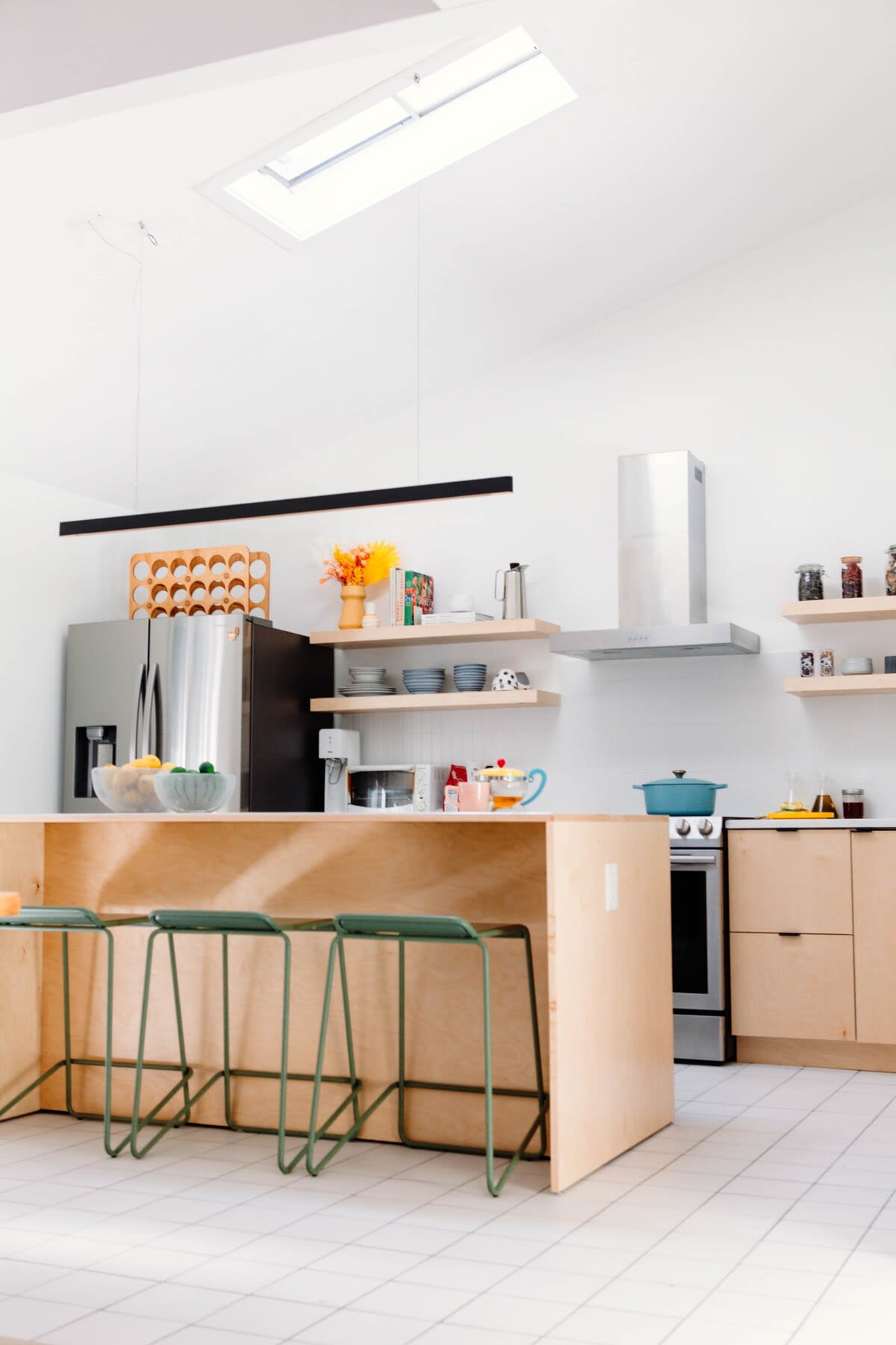 A modern kitchen features light wood cabinetry, a large island with green bar stools, and stainless steel appliances. Open shelving displays dishes and decor, with a skylight providing overhead natural light.