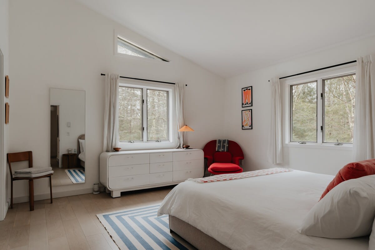 Bright bedroom with white walls and light wood floors, featuring a large bed, two windows overlooking a wooded landscape, a white dresser, and a vibrant red armchair. A full-length mirror and a blue and white striped rug complete the space.
