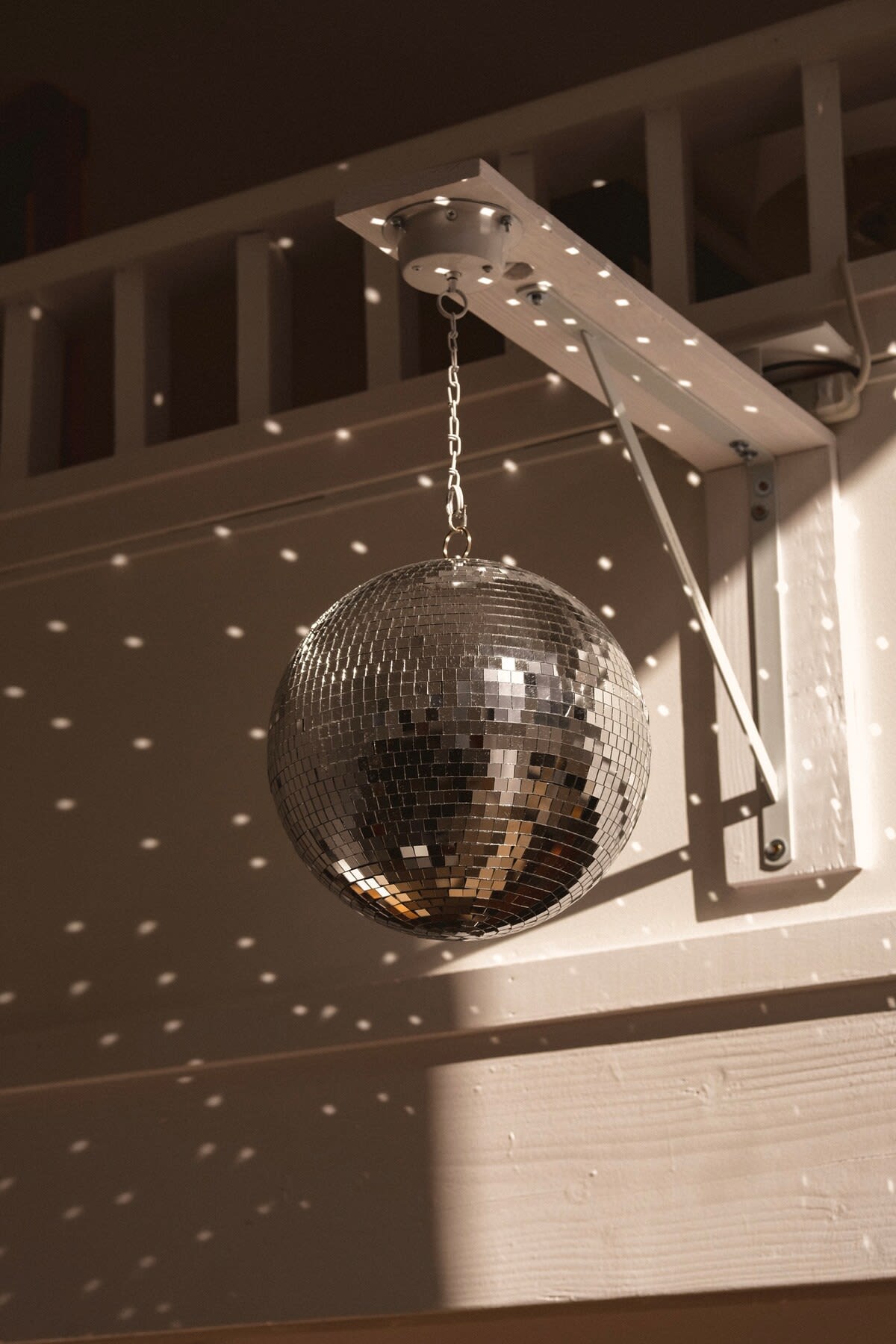 A close-up of a silver disco ball hanging from a white wooden bracket, casting numerous small, bright reflections onto the surrounding walls and ceiling.