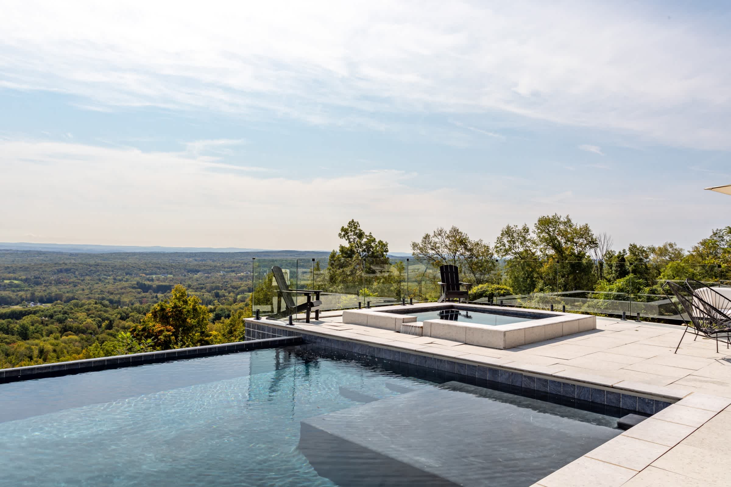 The infinity pool and adjacent hot tub are showcased with a clear glass railing, offering unobstructed views of the expansive forested valley below. Two dark Adirondack chairs face the vista.