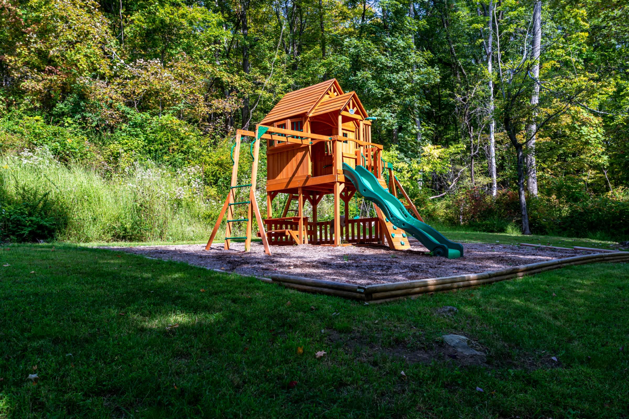 A wooden children's playset with a slide and swings is nestled in a grassy clearing, surrounded by lush green trees. The play area is bordered by a wooden retaining wall.