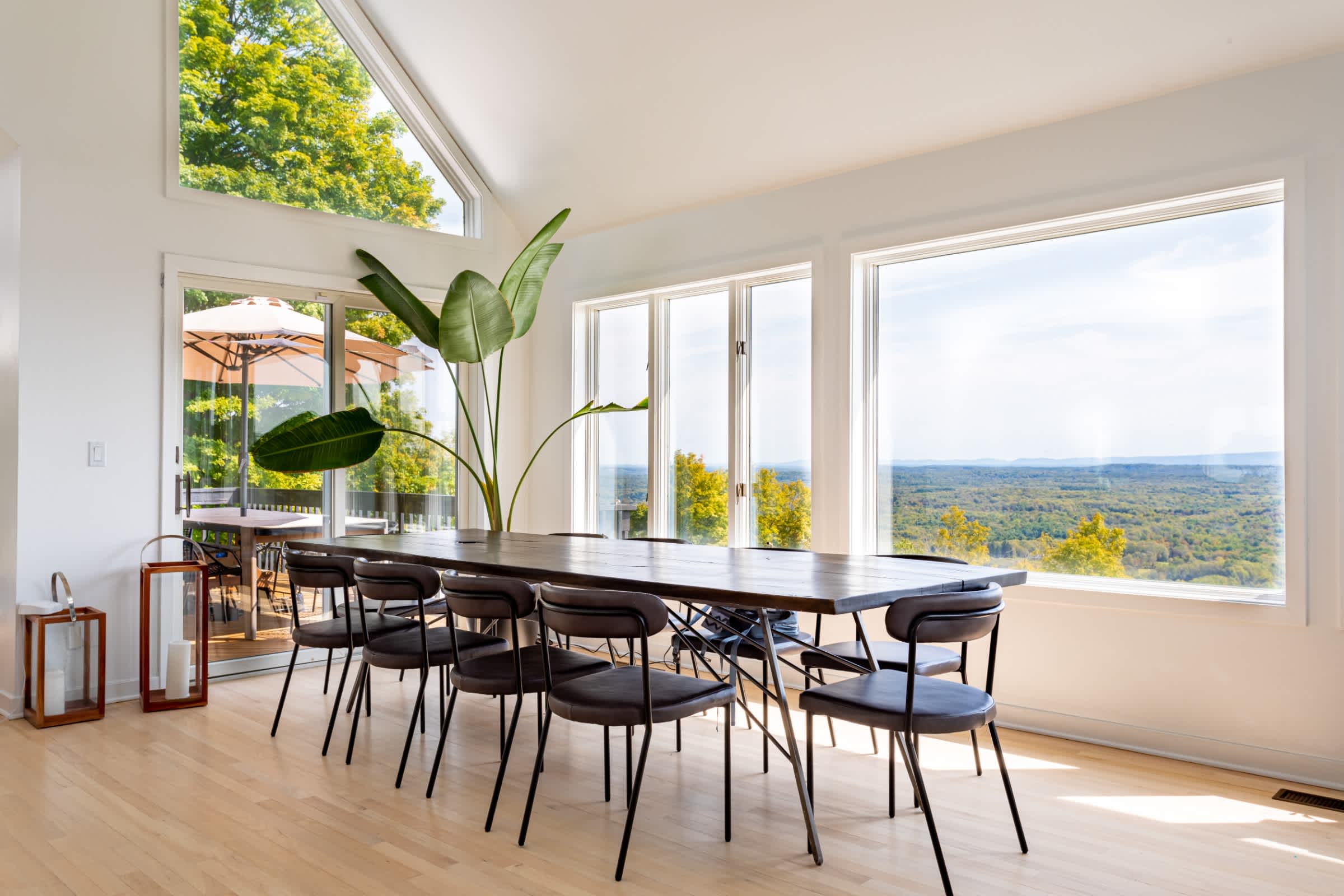 Spacious dining room featuring a long dark wood table with eight modern chairs, surrounded by large windows that provide panoramic views of the lush green landscape.