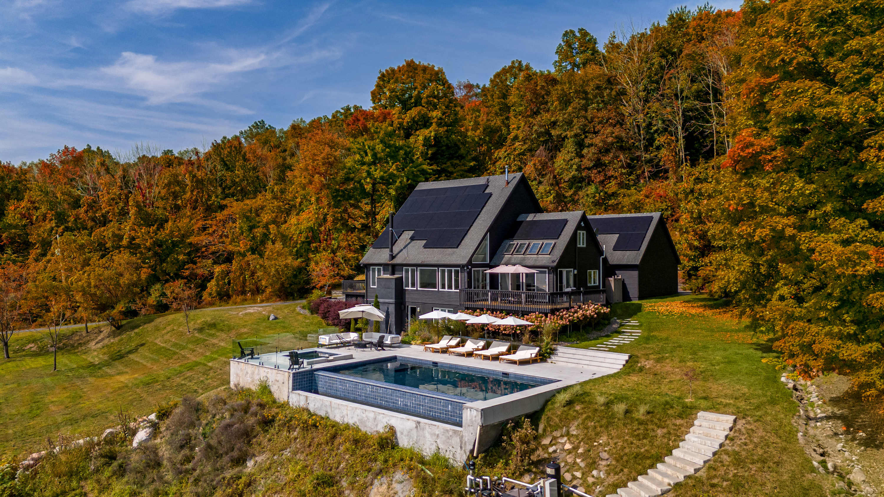 Aerial view of the modern dark-sided house with extensive solar panels on its roof, situated on a hillside. The infinity pool and patio are visible, surrounded by vibrant autumn foliage and distant mountain views.