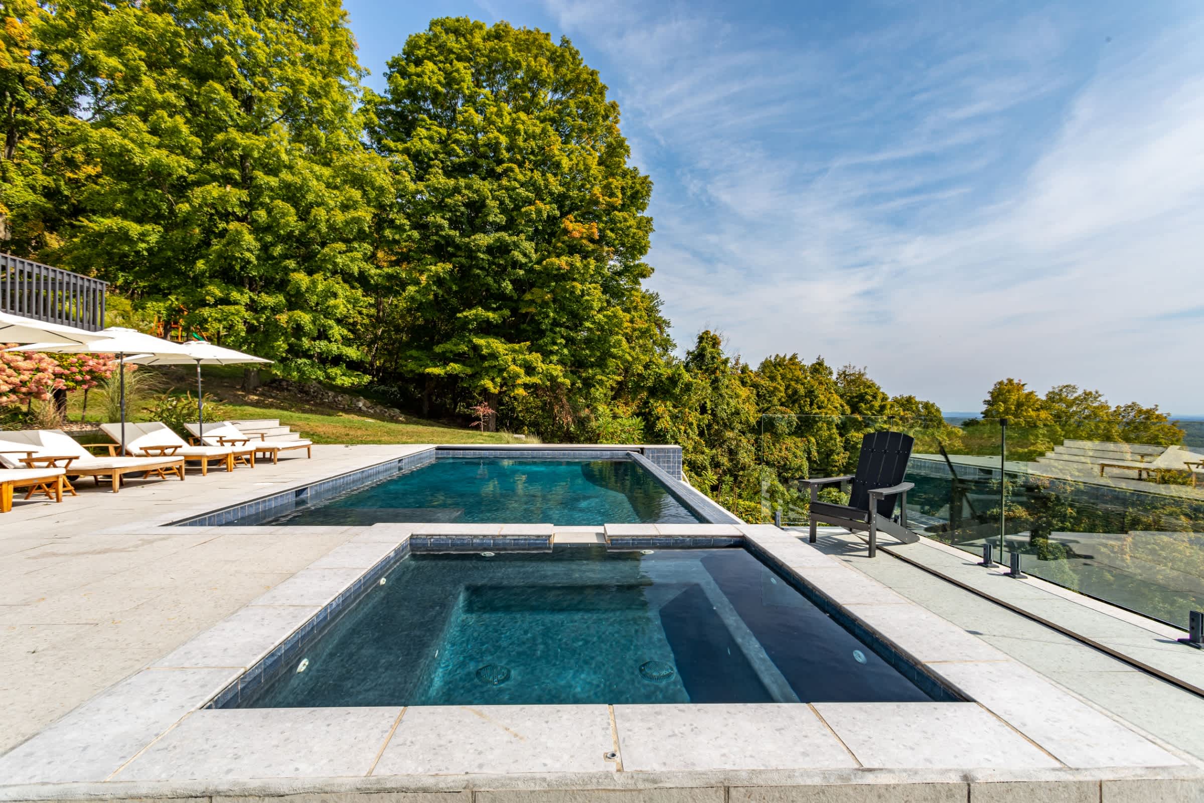 A closer view of the rectangular infinity pool and square hot tub, both featuring dark blue tiling and surrounded by a light-colored stone patio. A glass railing ensures clear mountain views.