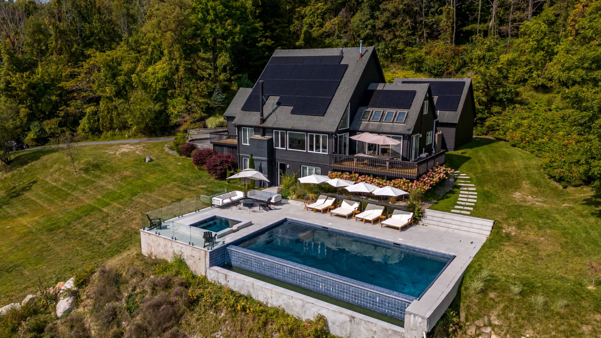 Aerial view of a modern dark-sided house with solar panels on the roof, featuring an infinity pool and hot tub on a patio, set on a hillside surrounded by lush green forests.