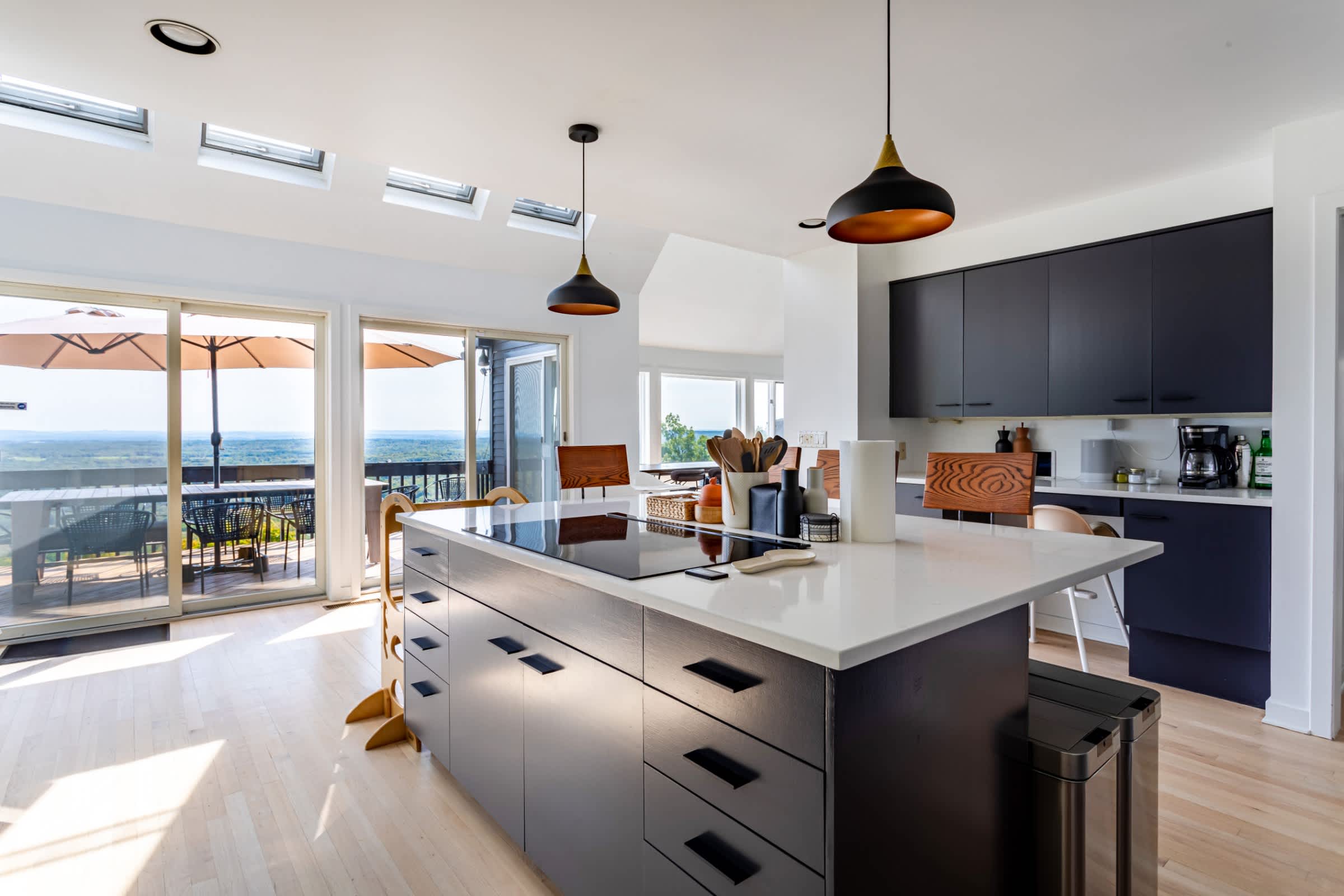 Modern kitchen with dark flat-panel cabinetry, a large central island with a white countertop and integrated cooktop, pendant lights, and skylights, with sliding doors leading to an outdoor deck.