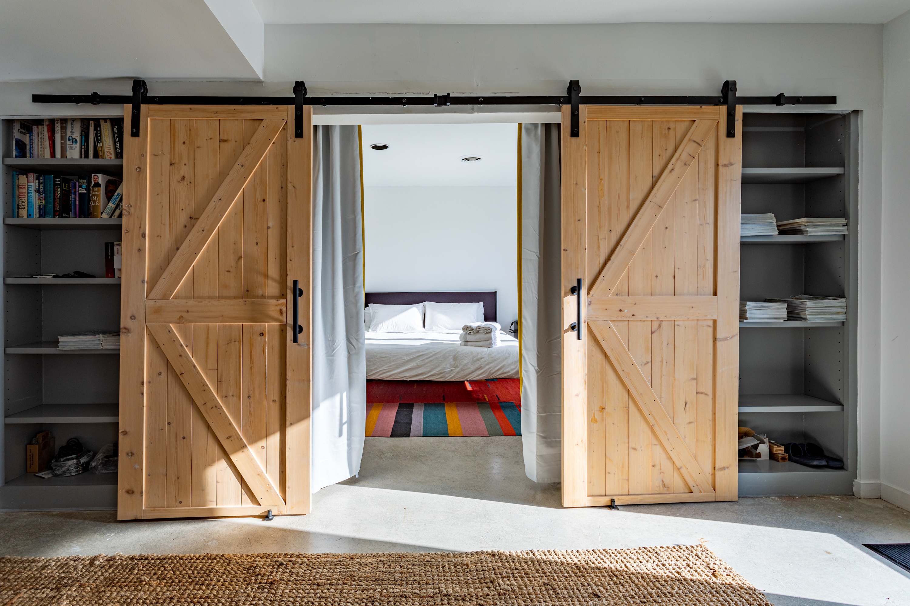 A view of two large wooden barn doors opening to reveal a glimpse of a bedroom with a colorful striped rug, flanked by built-in shelving units filled with books and decor.