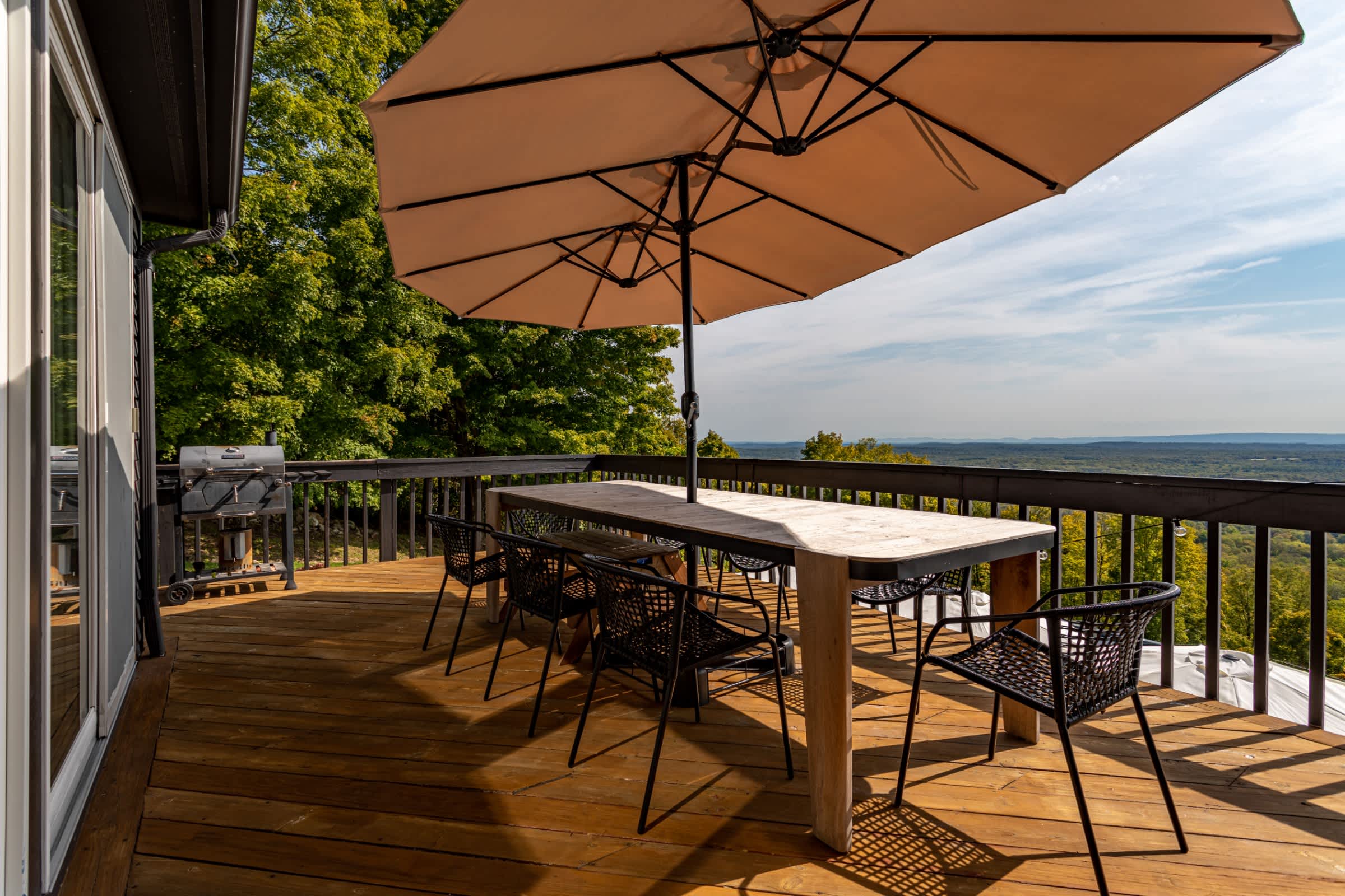 Spacious wooden deck furnished with a large outdoor dining table, black chairs, and a patio umbrella, offering a stunning panoramic view of a lush green forest and distant mountains.