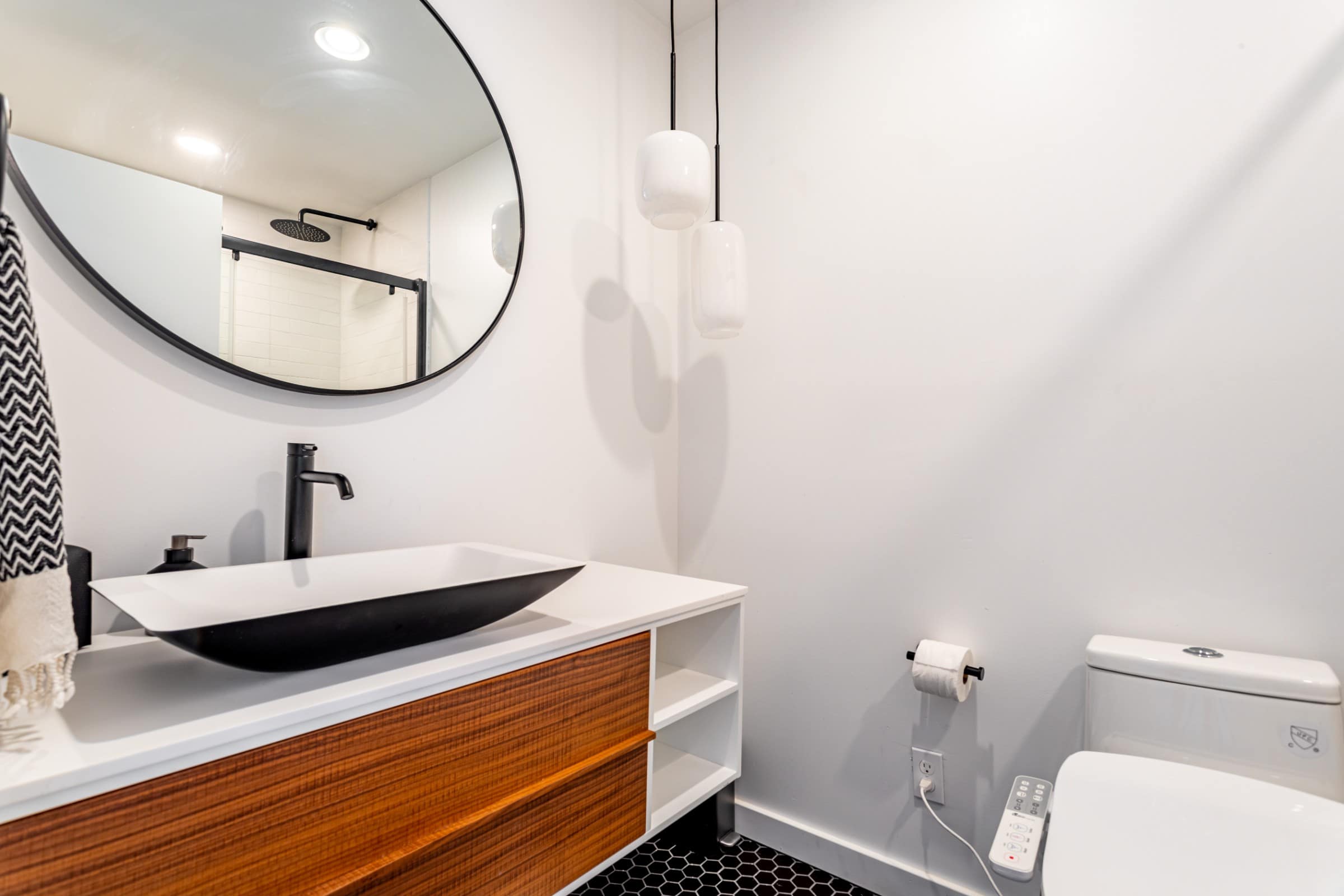 A sleek bathroom featuring a unique black and white vessel sink on a wooden vanity, a large round mirror, modern pendant lights, and a shower with black hexagonal floor tiles.