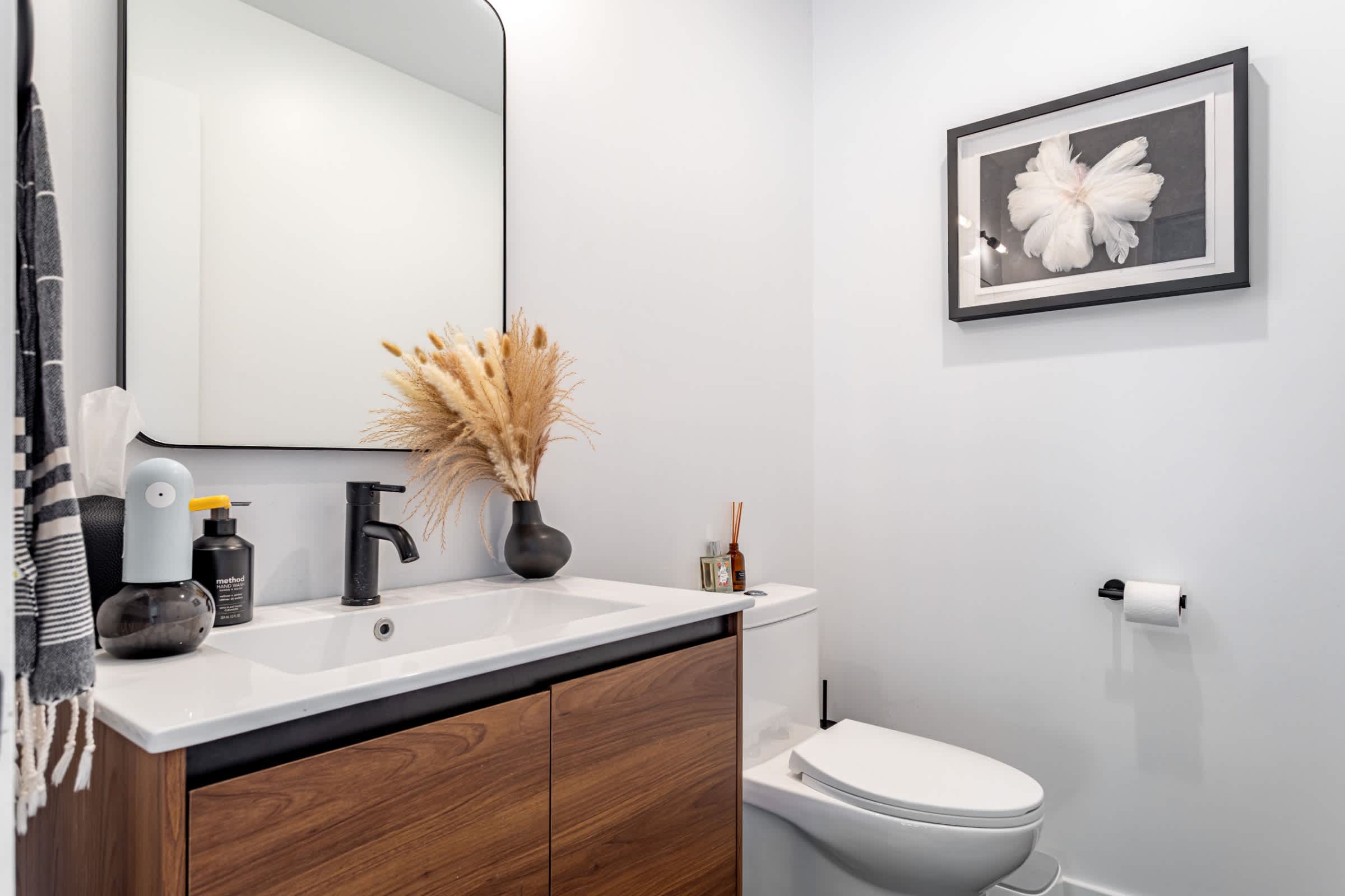 Modern powder room with a floating wooden vanity, white sink, black faucet, and a large rectangular mirror, featuring white walls and a framed floral art print.