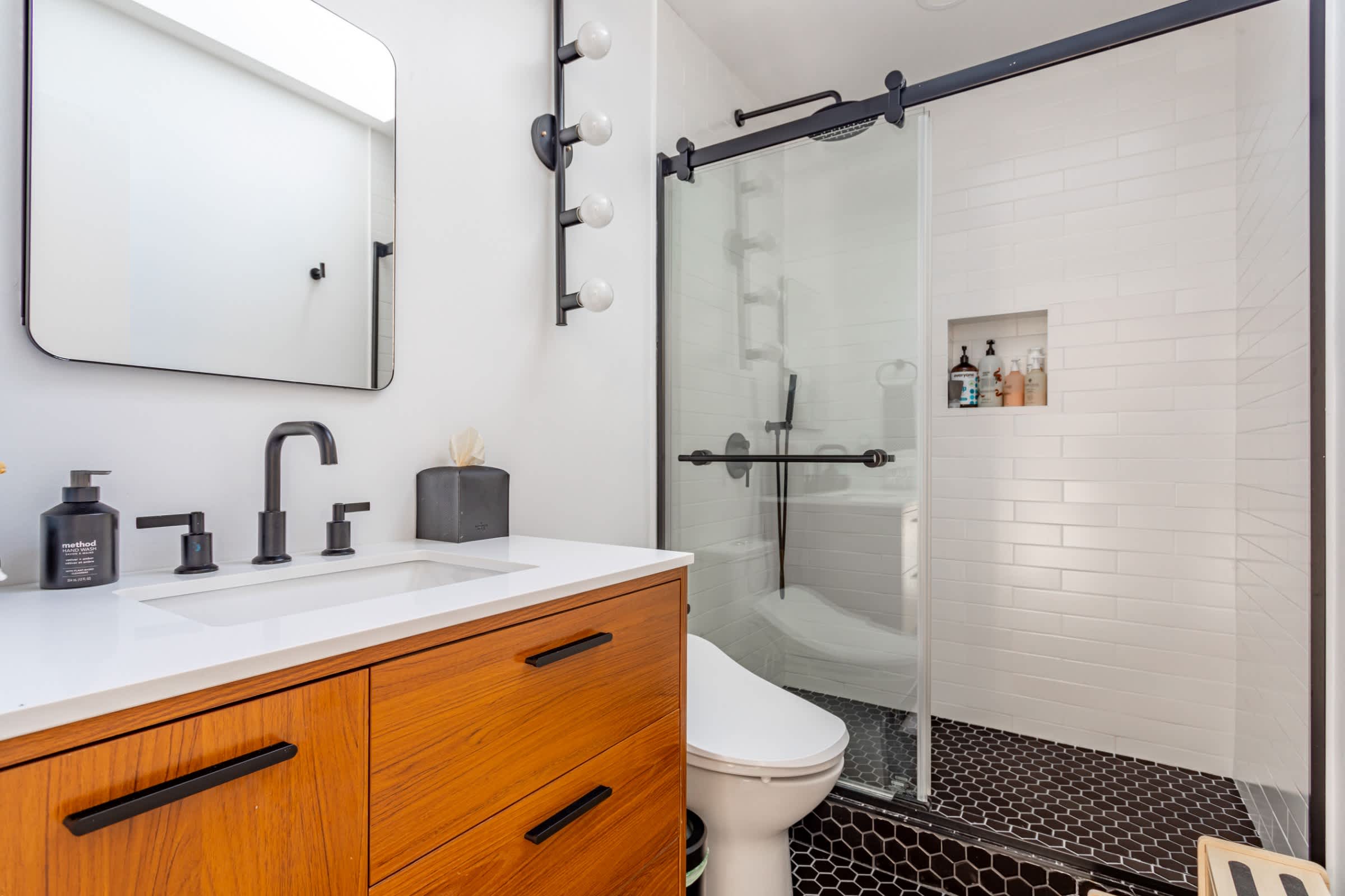 A modern bathroom showcasing a wooden vanity with a white countertop and black faucet, a rectangular mirror, and a walk-in shower with black hexagonal floor tiles and white subway wall tiles.