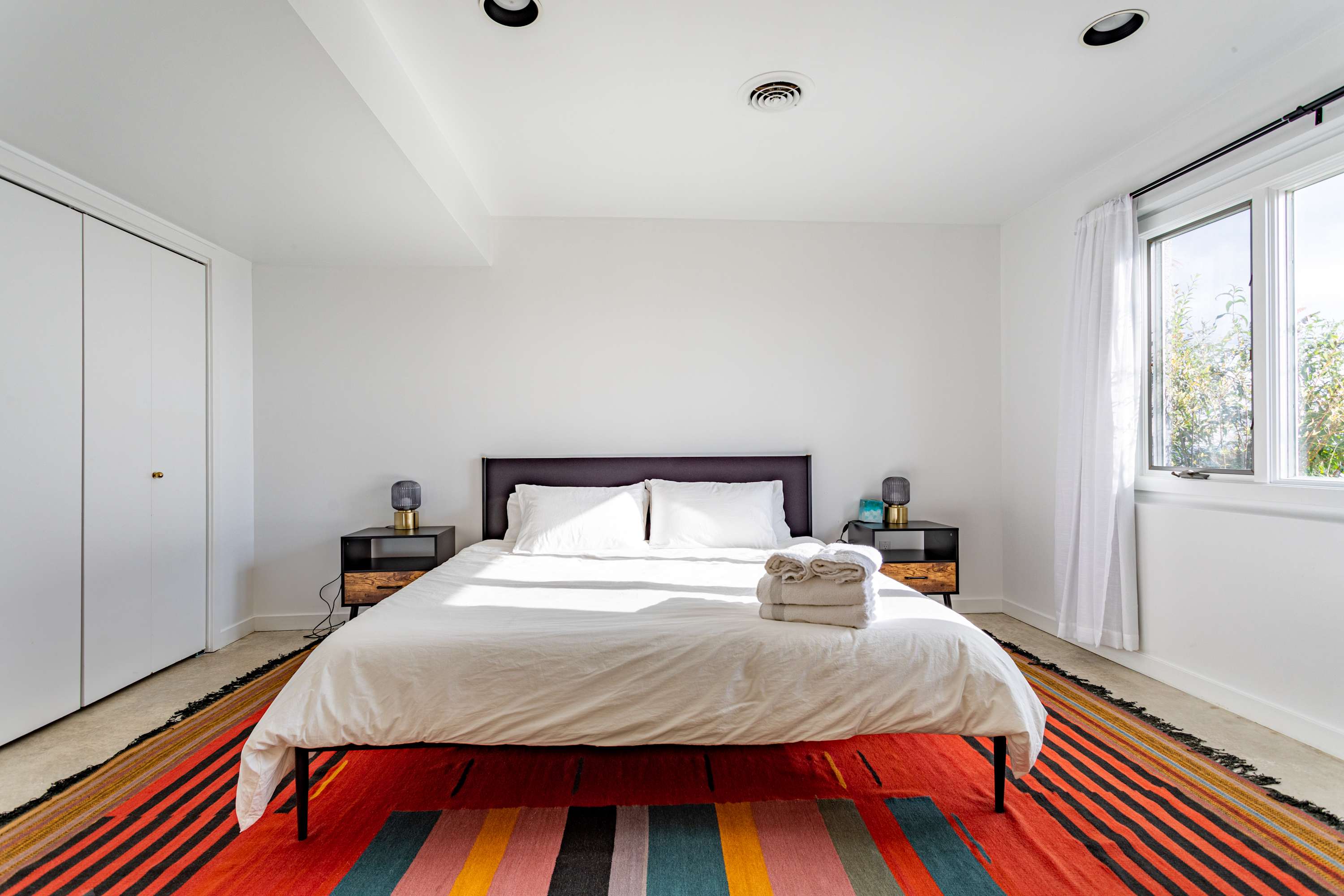 A bright bedroom featuring a queen-sized bed with a dark upholstered headboard, two modern nightstands with lamps, and a vibrant striped rug in red, orange, and blue tones.