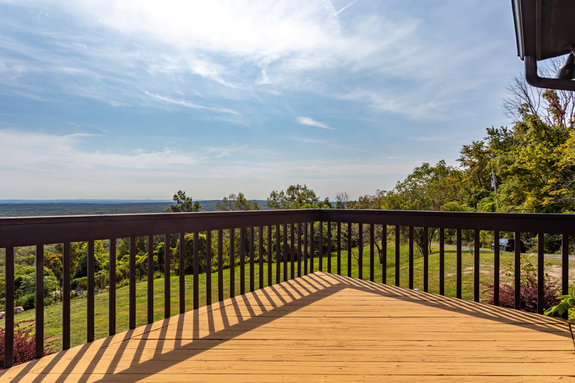 Expansive view from a wooden deck with dark railings, showcasing a vast landscape of rolling green hills and distant forests under a bright blue sky with scattered clouds.