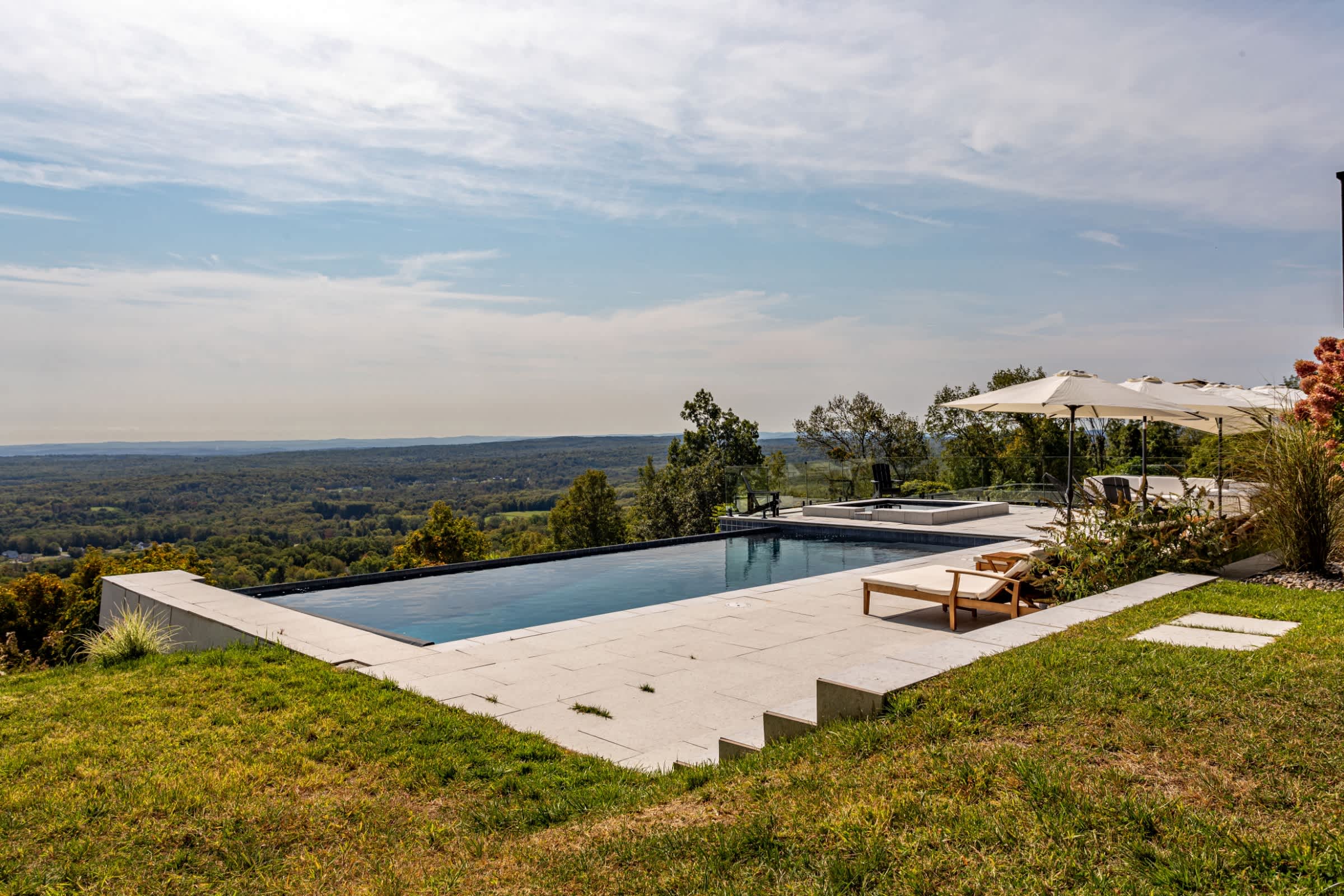 Infinity-edge swimming pool with a light-colored stone patio and lounge chairs, situated on a hillside with panoramic views of a vast, tree-covered valley under a bright sky.