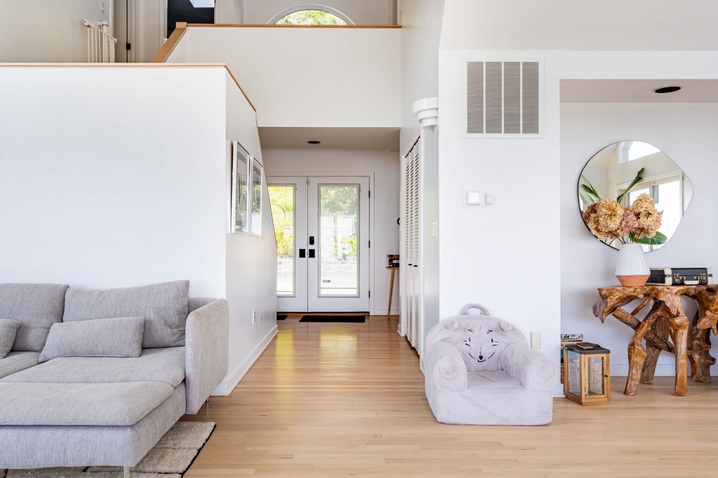 Bright and airy foyer with light hardwood floors, white walls, a grey sectional sofa, and a unique wooden console table with a round mirror, leading to an open living space.