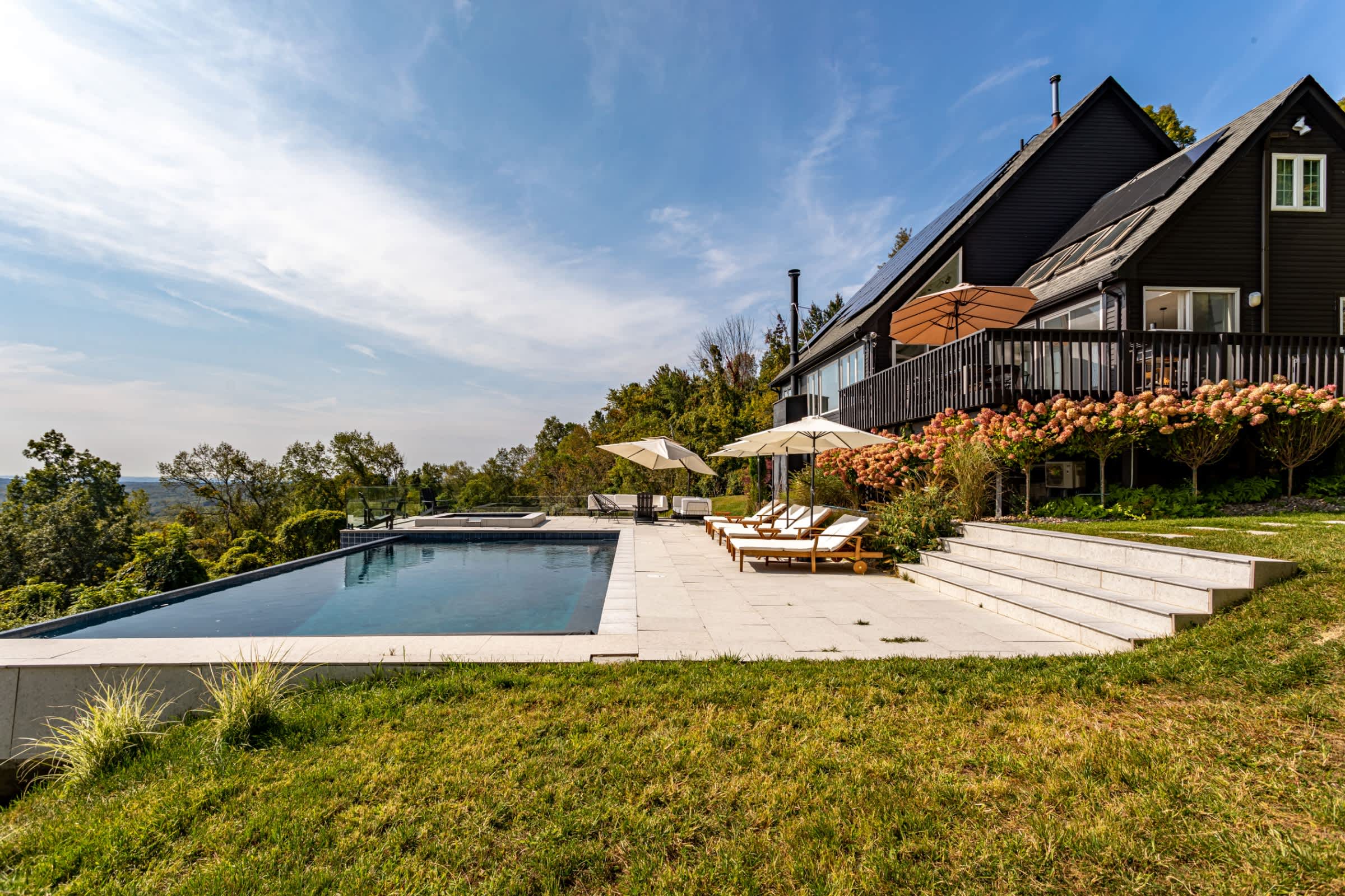 Expansive infinity-edge pool and integrated hot tub on a stone patio, overlooking a vast mountain landscape. The modern dark-sided house with solar panels and a deck is visible in the background.