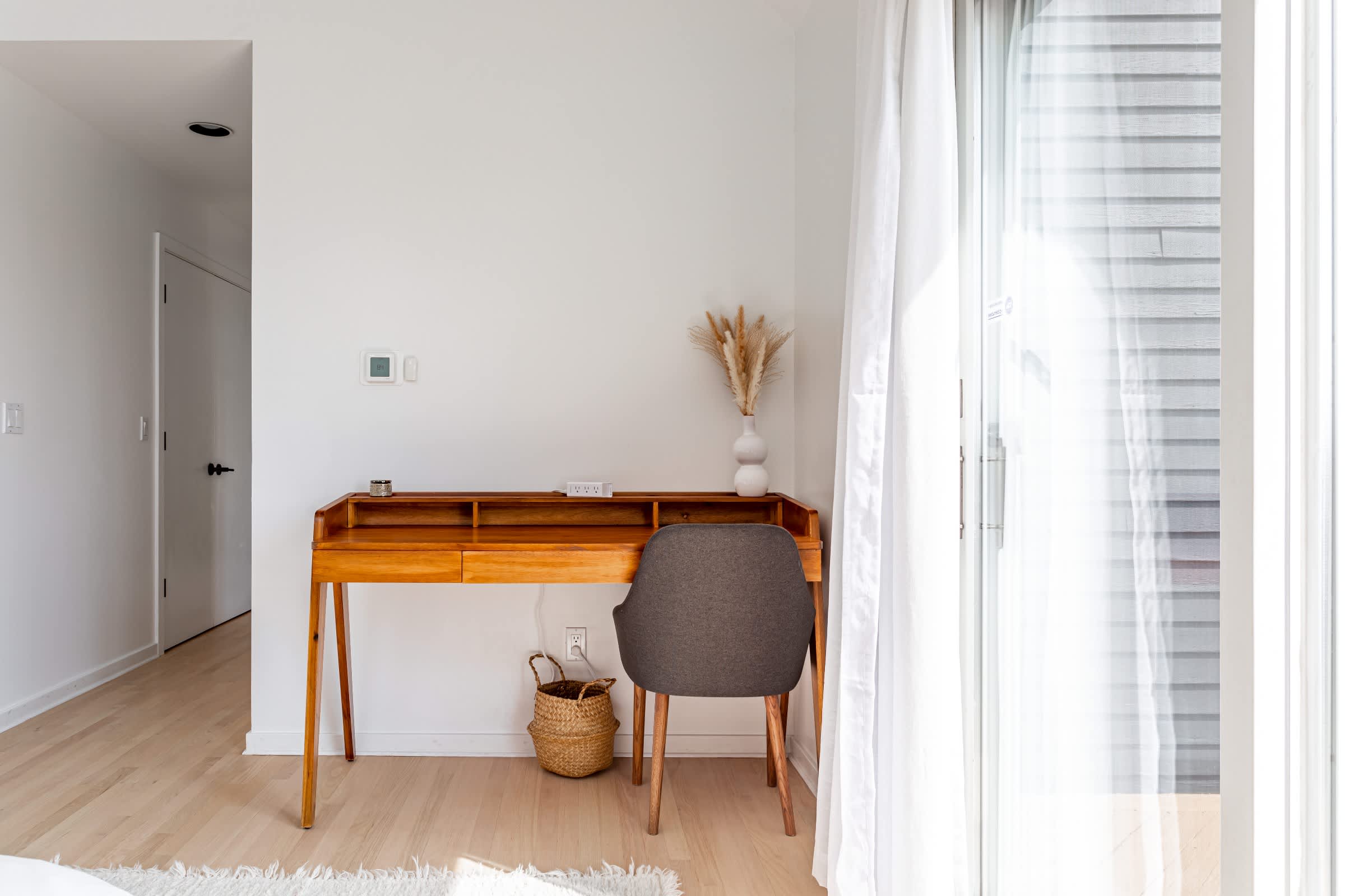 Dedicated workspace within the bedroom featuring a mid-century modern wooden desk and a grey upholstered chair, positioned next to sliding glass doors for natural light.