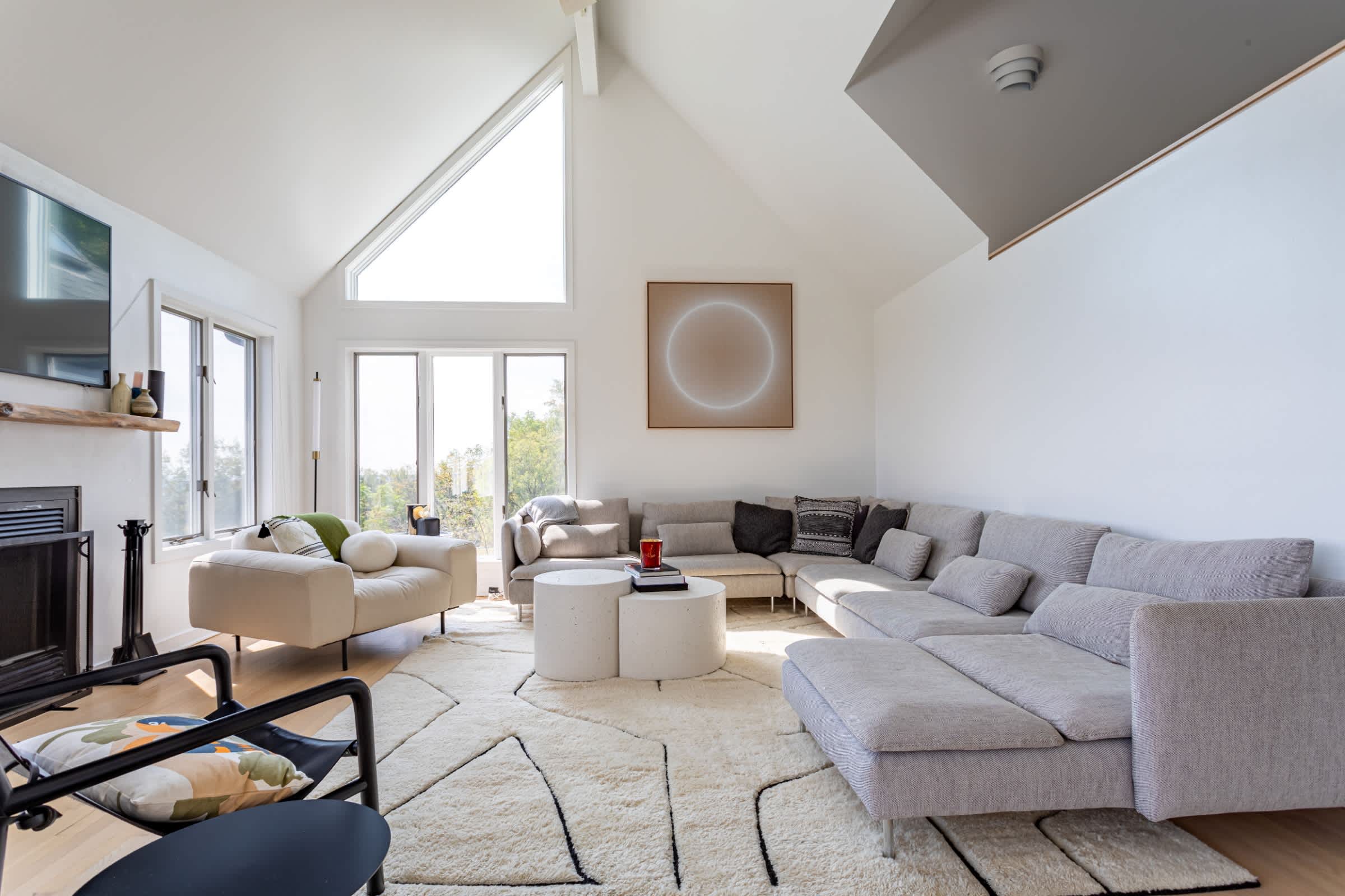 Contemporary living room with high vaulted ceilings and large triangular windows, featuring a spacious sectional sofa, an armchair, and two round concrete coffee tables on a geometric area rug.