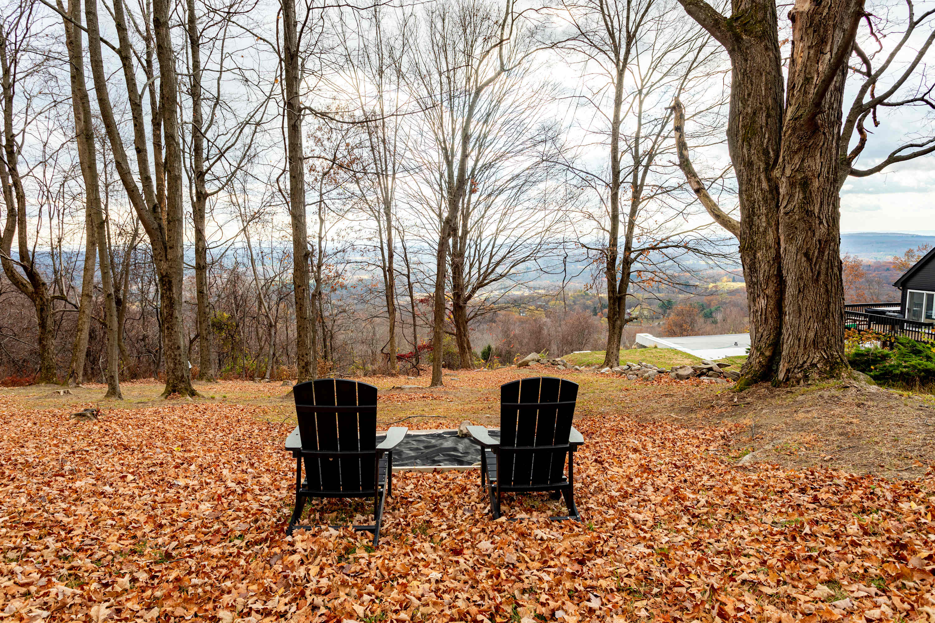 Two black Adirondack chairs and a small table are set on a carpet of fallen autumn leaves, offering a serene spot to enjoy the distant valley views through bare trees.