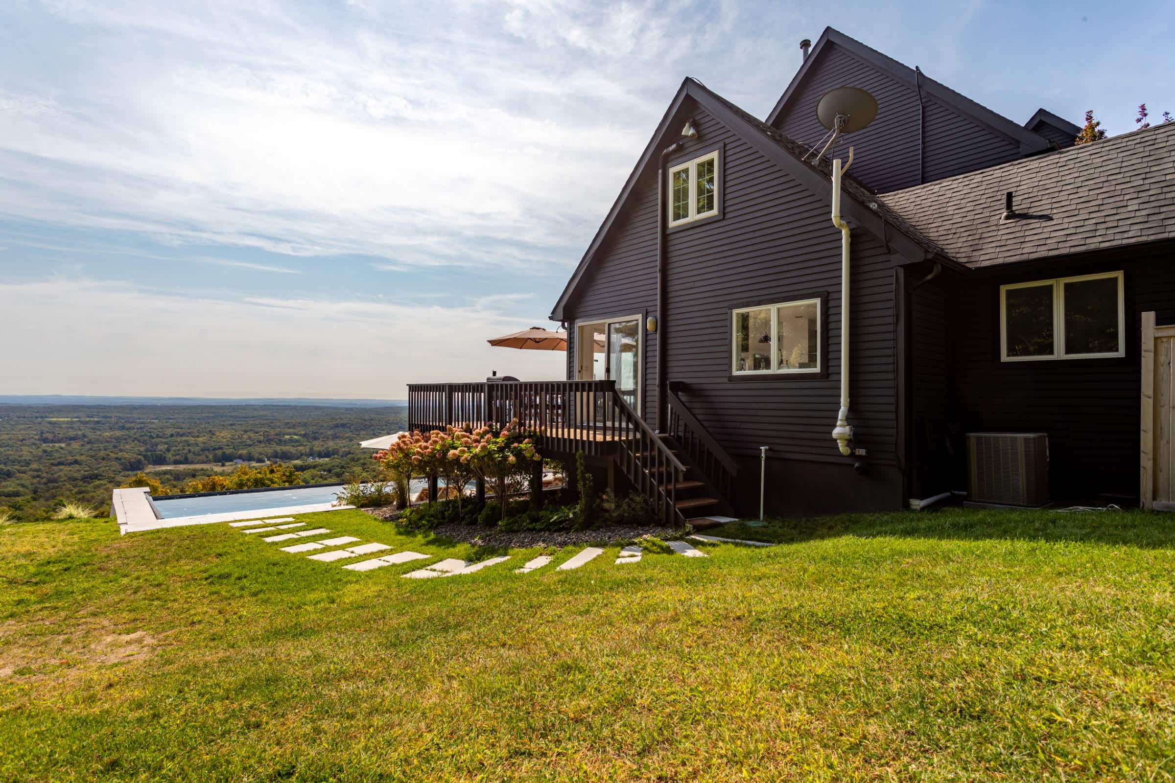 Modern dark-sided house with a large wooden deck and an infinity-edge pool, offering expansive panoramic views of a lush green valley under a partly cloudy sky.