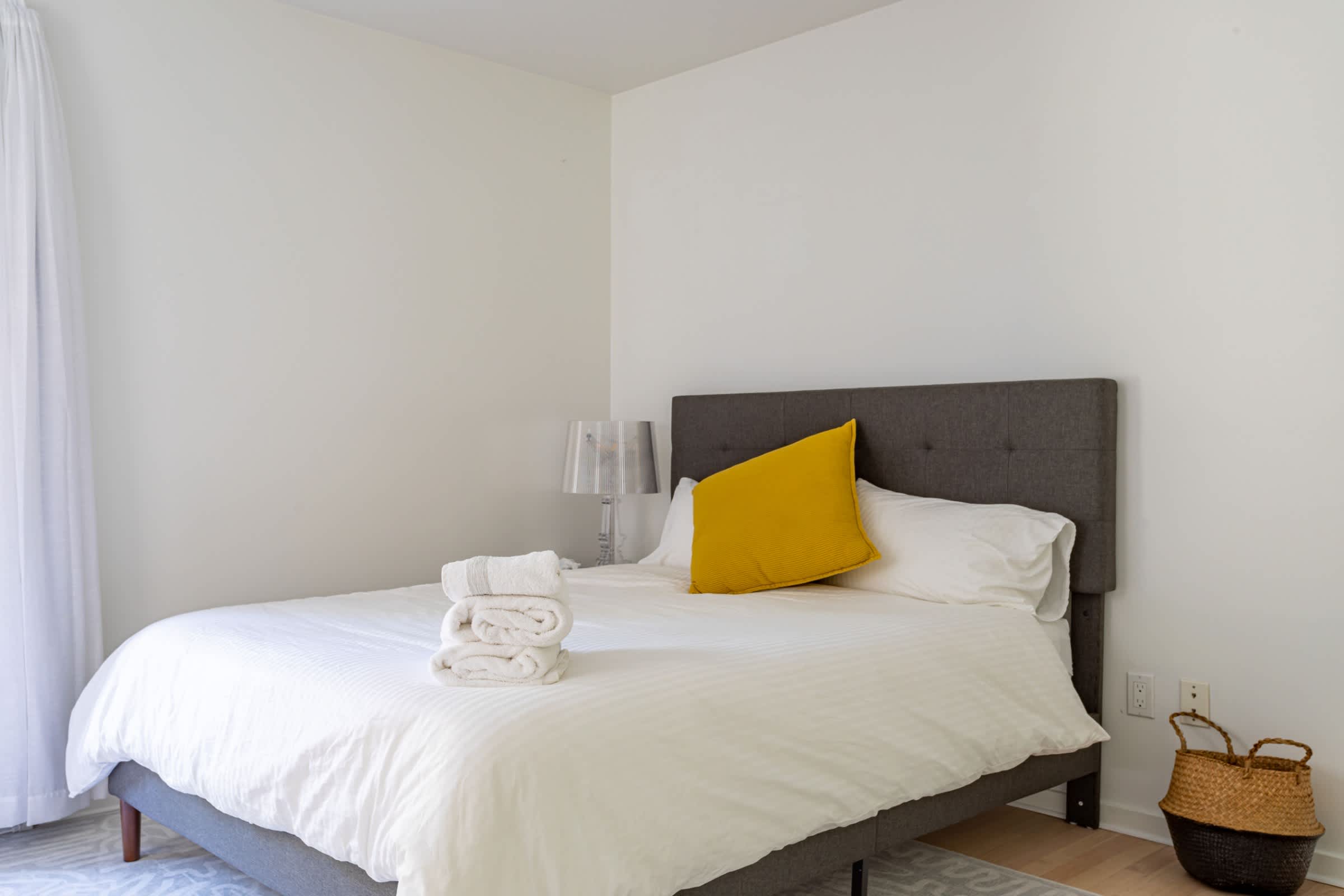 A bright and minimalist bedroom featuring a gray upholstered bed with white bedding and a vibrant yellow throw pillow, complemented by a glass-based lamp on a simple nightstand.