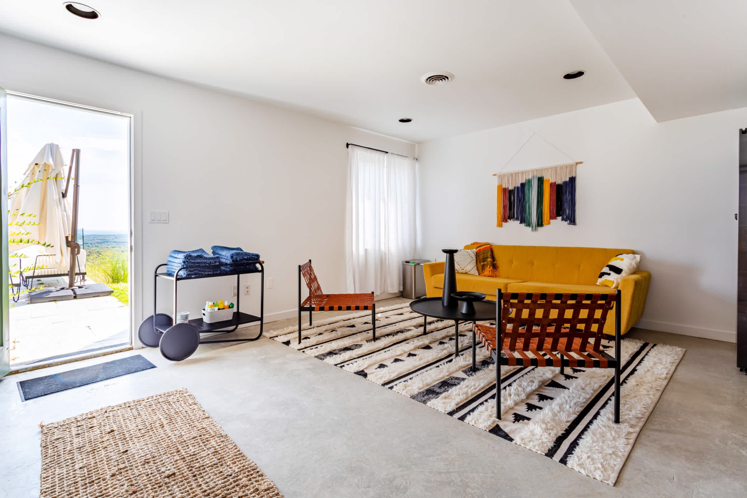 A vibrant living area with a bright yellow sofa, two woven leather chairs, a black round coffee table, and a geometric area rug, with an open door leading to an outdoor space.