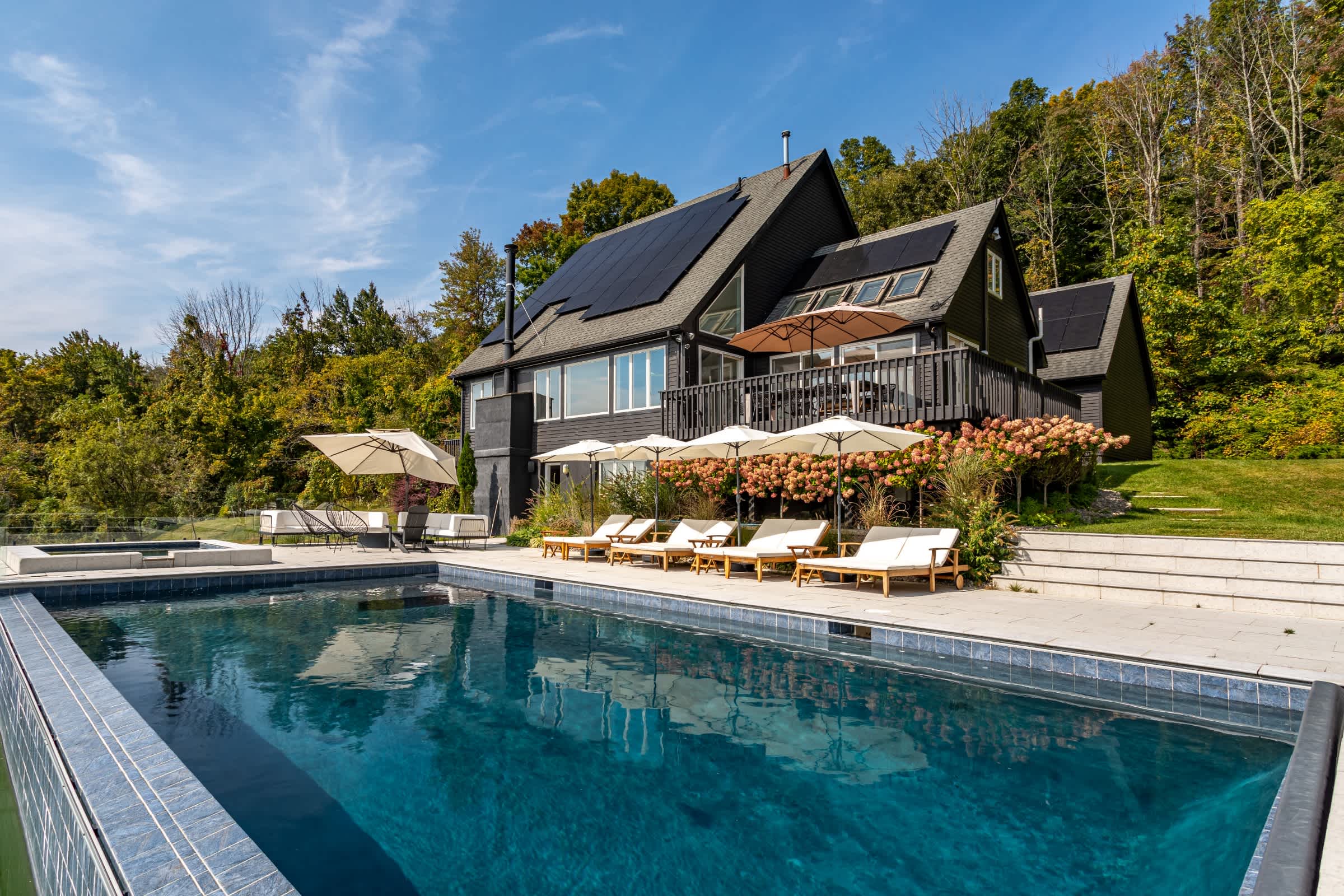 Wide view of the dark-sided house with solar panels, featuring a large infinity pool and hot tub on a stone patio. Lush trees and distant mountains frame the scenic outdoor living space.