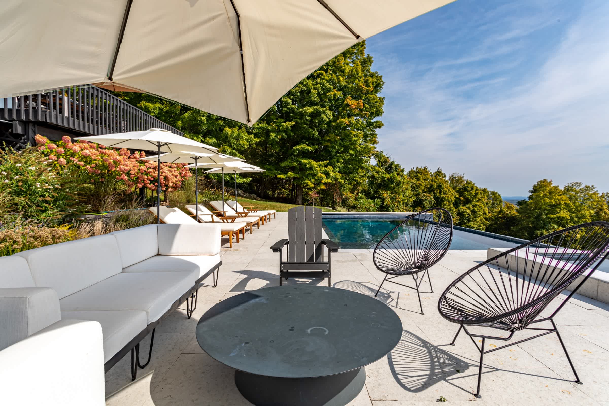 Outdoor lounge area on a stone patio, featuring a white sectional sofa, black wire chairs, and a round coffee table under a large umbrella. The infinity pool is visible in the background.