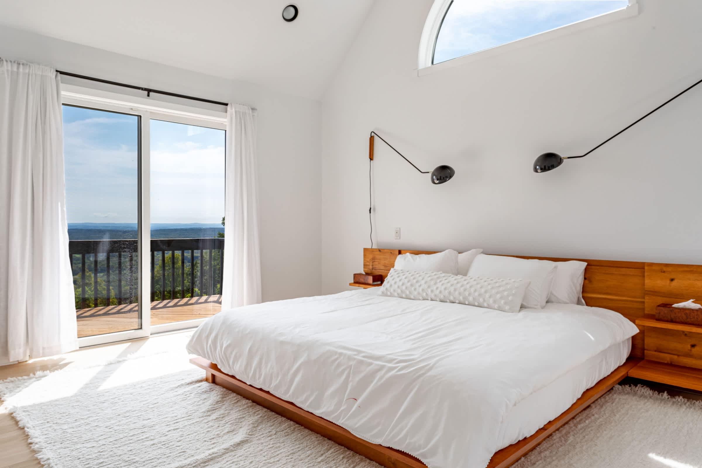 Serene bedroom with a king-sized bed, custom wooden platform bed frame and headboard, white bedding, and sliding glass doors opening to a private deck with scenic views.