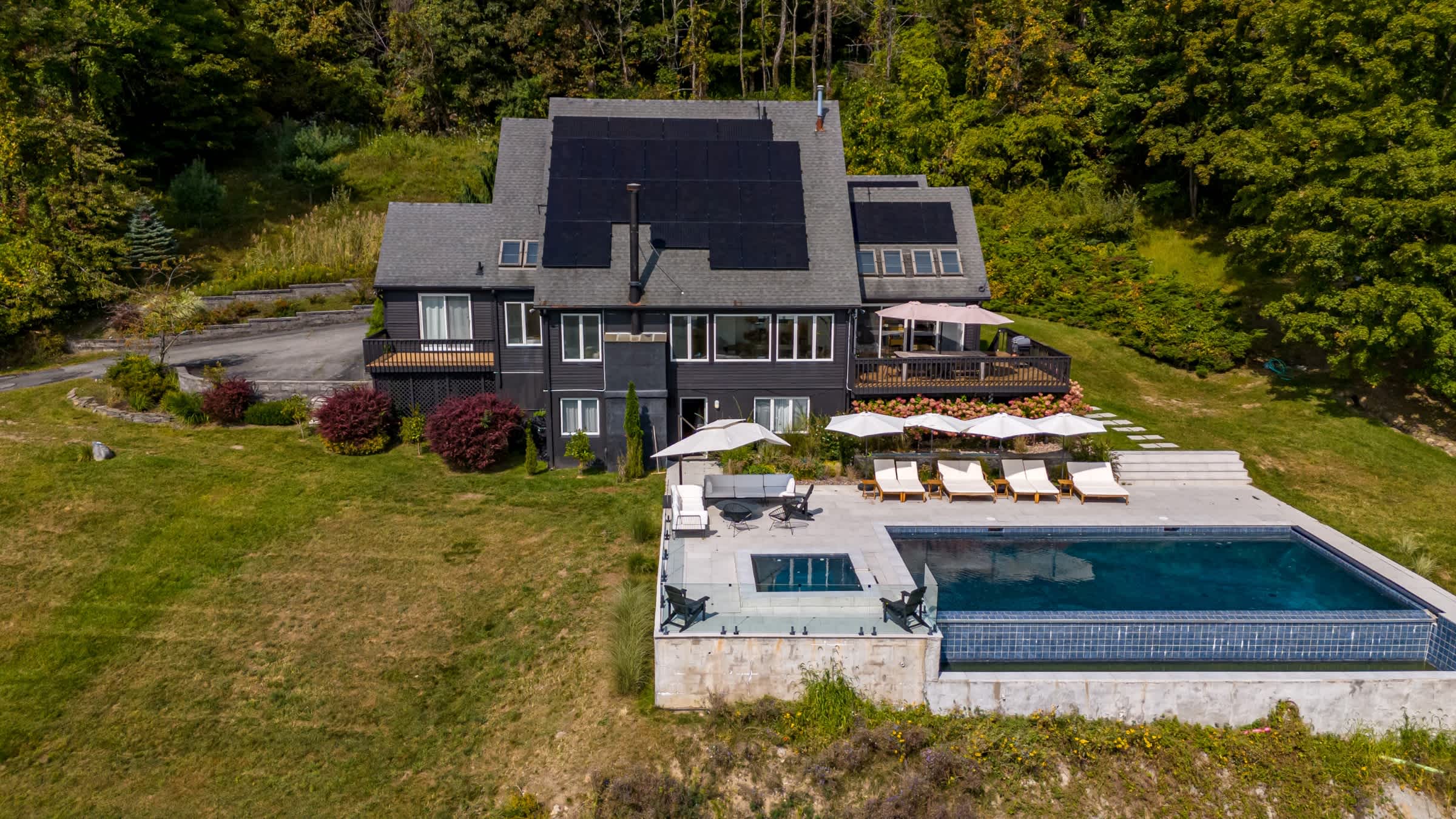 High aerial view showcasing the dark-colored house with a large array of solar panels on its roof, an infinity pool, and a spacious patio. The property is nestled amidst a lush, green landscape with distant views.