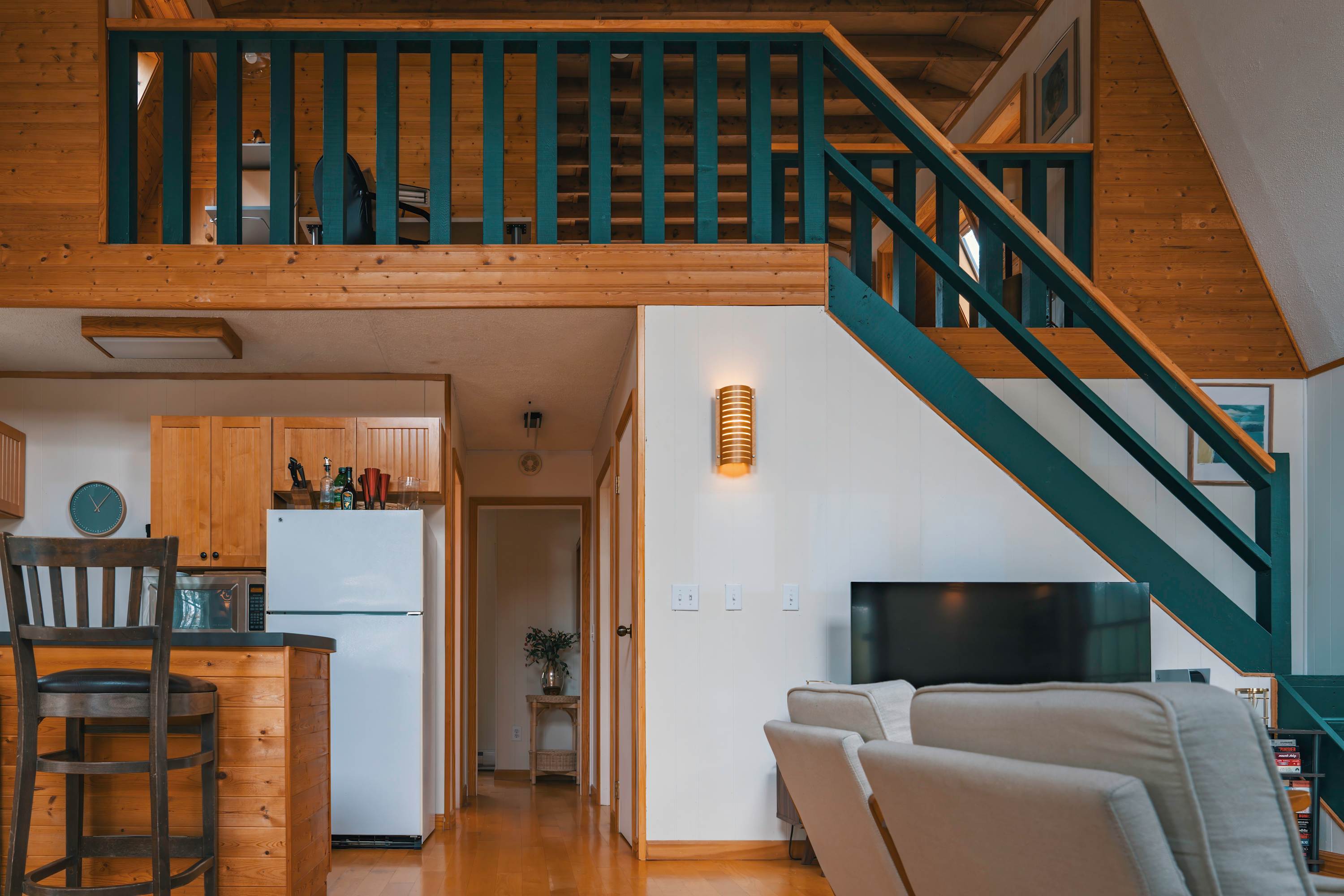 View of an open-concept living space with a dark teal wooden staircase and railing leading to an upper loft. A white sofa faces a large television, and a kitchen area with a breakfast bar is visible to the left, all set on warm hardwood floors.