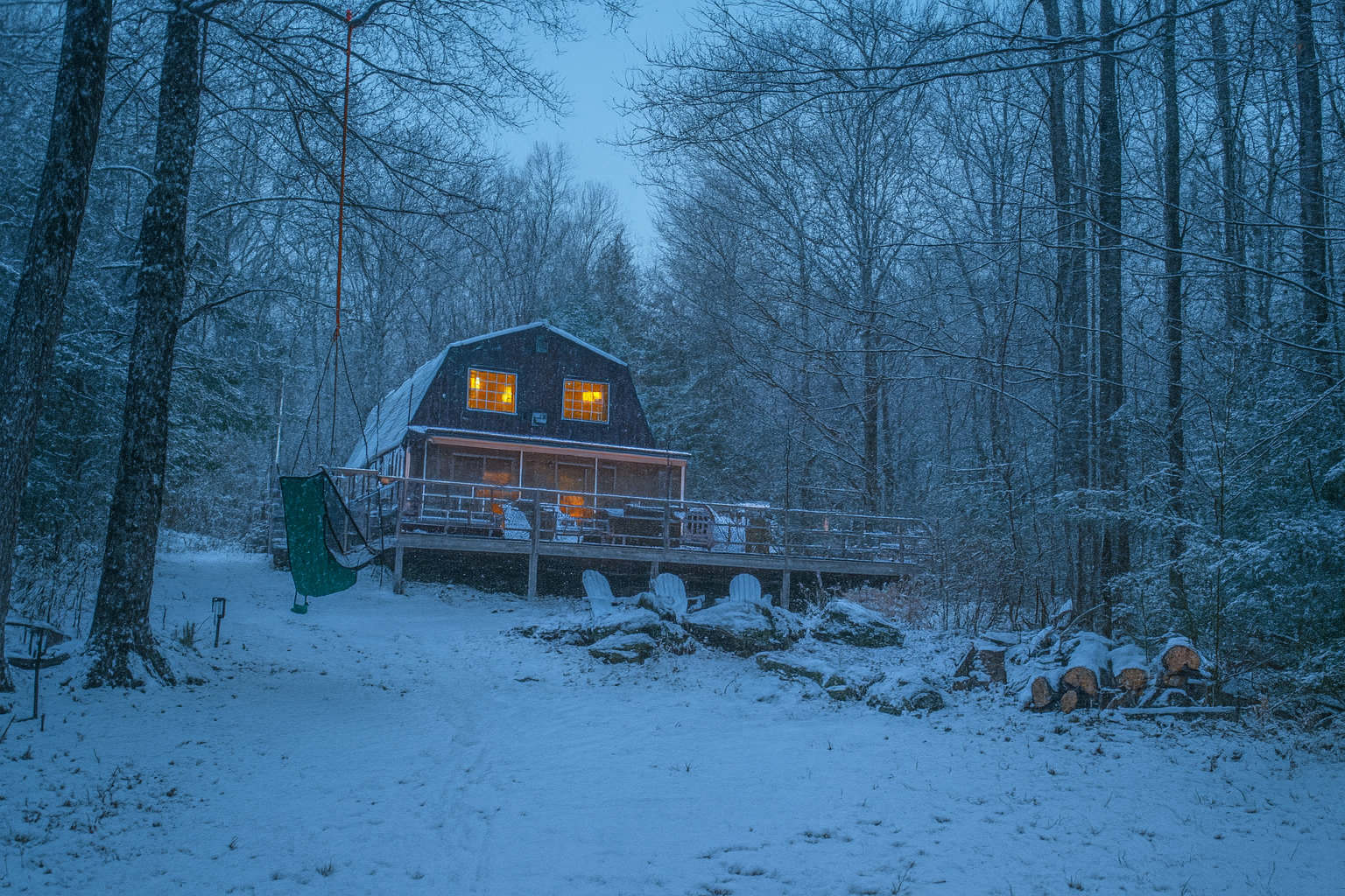 A cozy house with illuminated windows stands amidst a snow-covered forest under a soft blue twilight sky, featuring a large deck with white outdoor seating and a hanging green swing chair, with stacked firewood nearby.