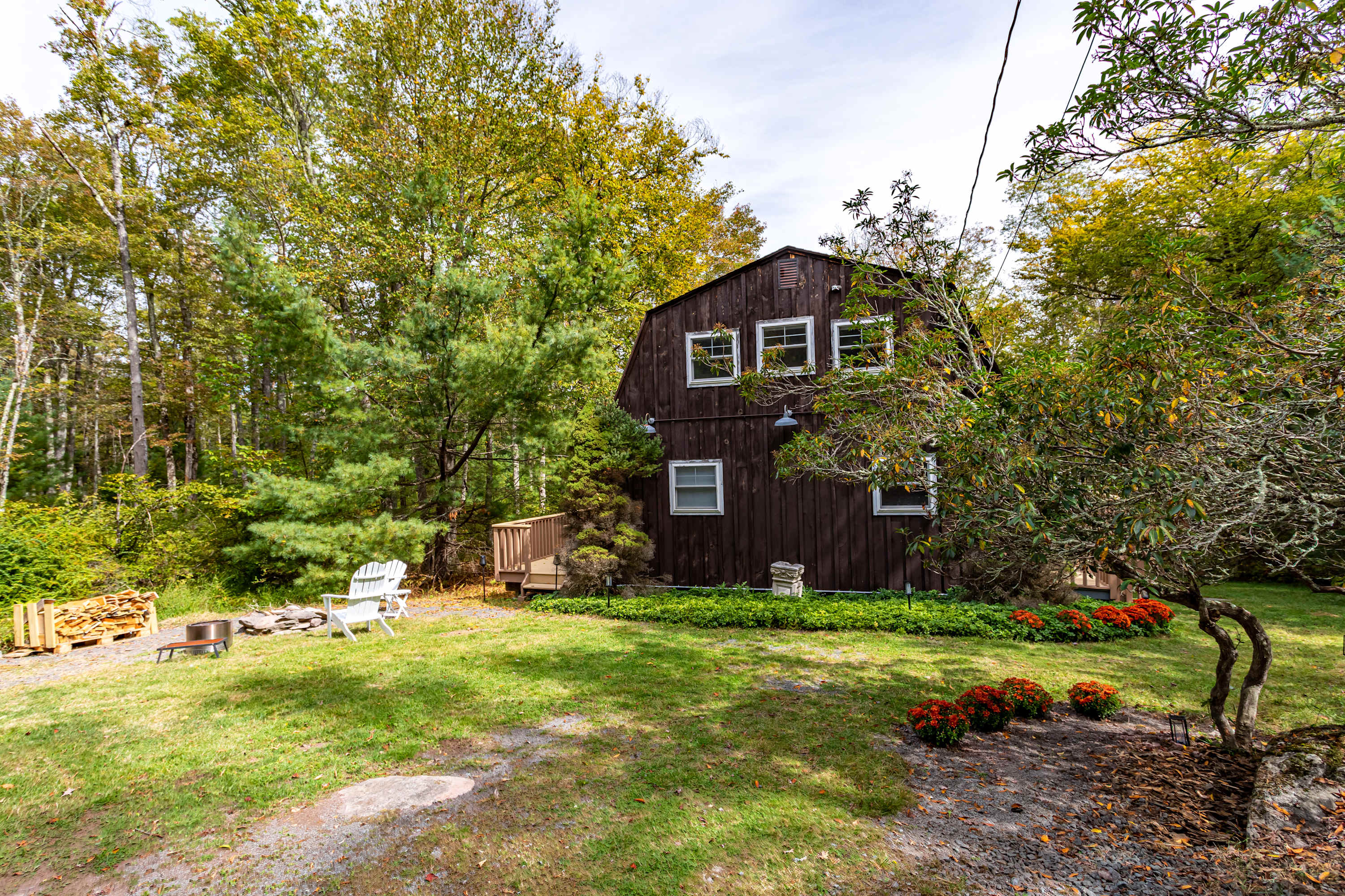 Front exterior view of a dark brown house with a barn-style roof, surrounded by lush green and autumn-colored trees. A small grassy yard features a fire pit and stacked firewood, with a white Adirondack chair, suggesting a cozy outdoor space.