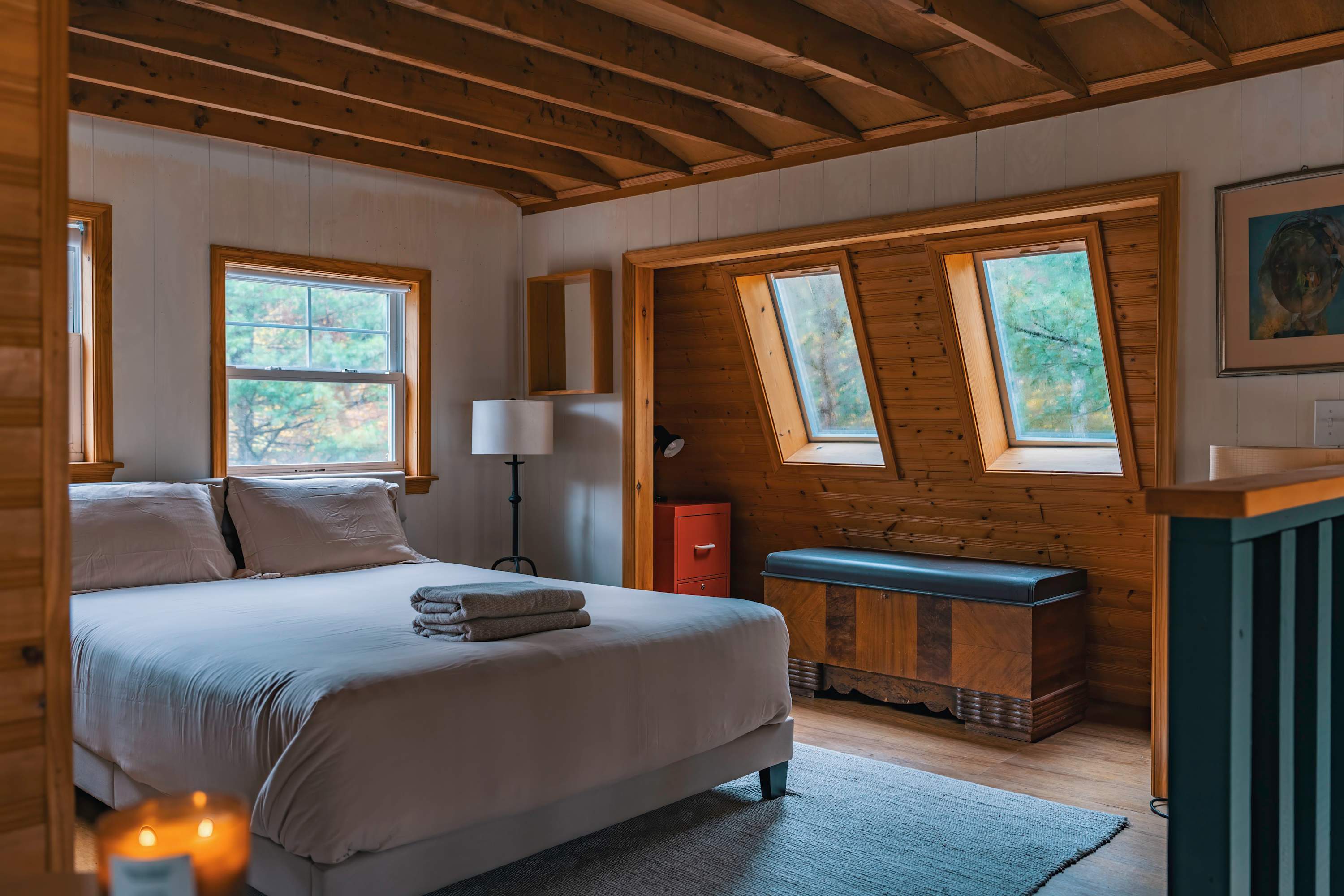 Cozy bedroom featuring a queen-sized bed with light bedding, exposed wooden beams on the ceiling, and white paneled walls. Two large windows with wooden frames offer views of the surrounding trees, complemented by a small red cabinet and a floor lamp.