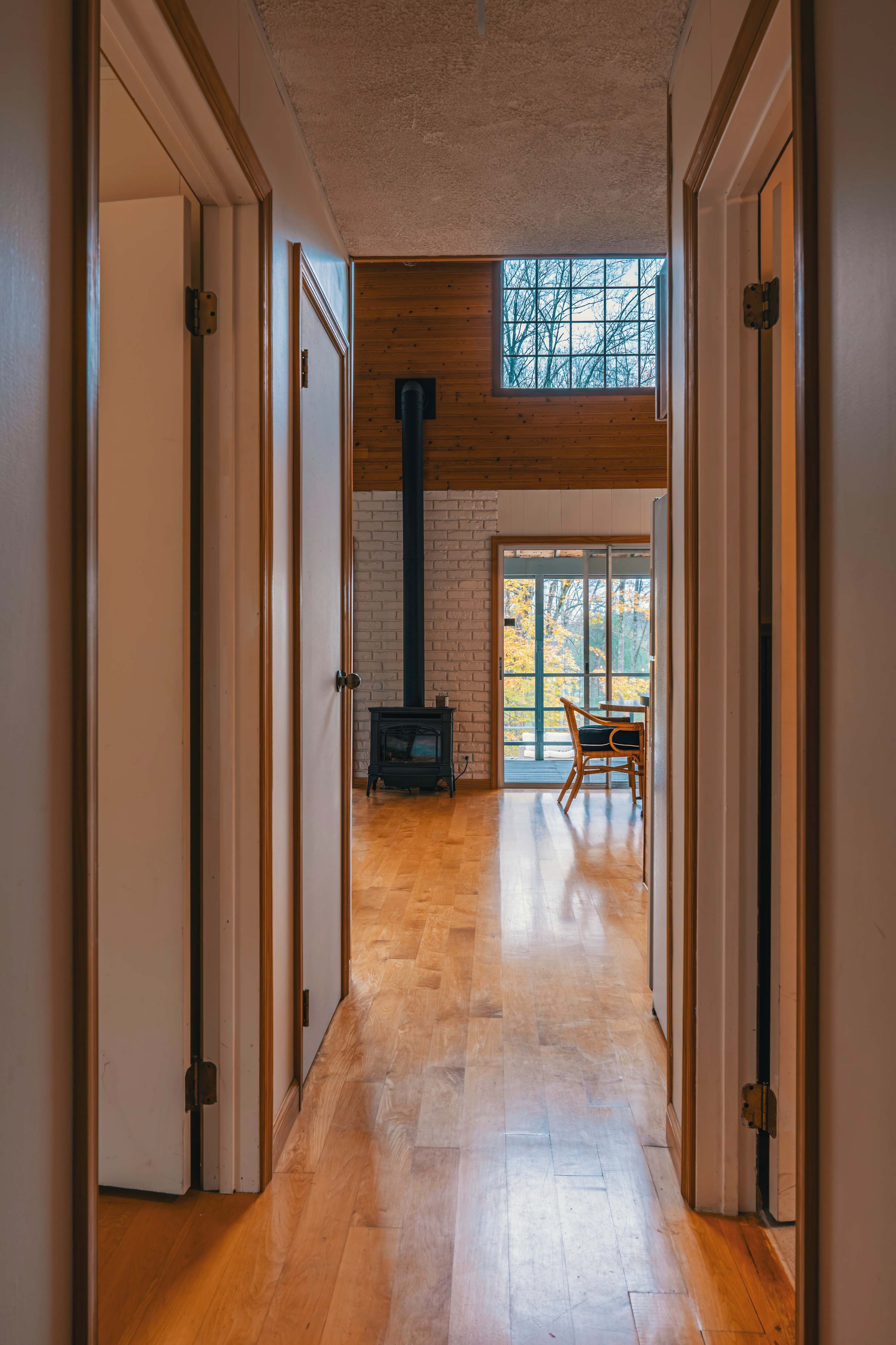 Narrow hallway with light-colored doors and polished hardwood floors, leading towards a bright living space with a black wood-burning stove and large windows.