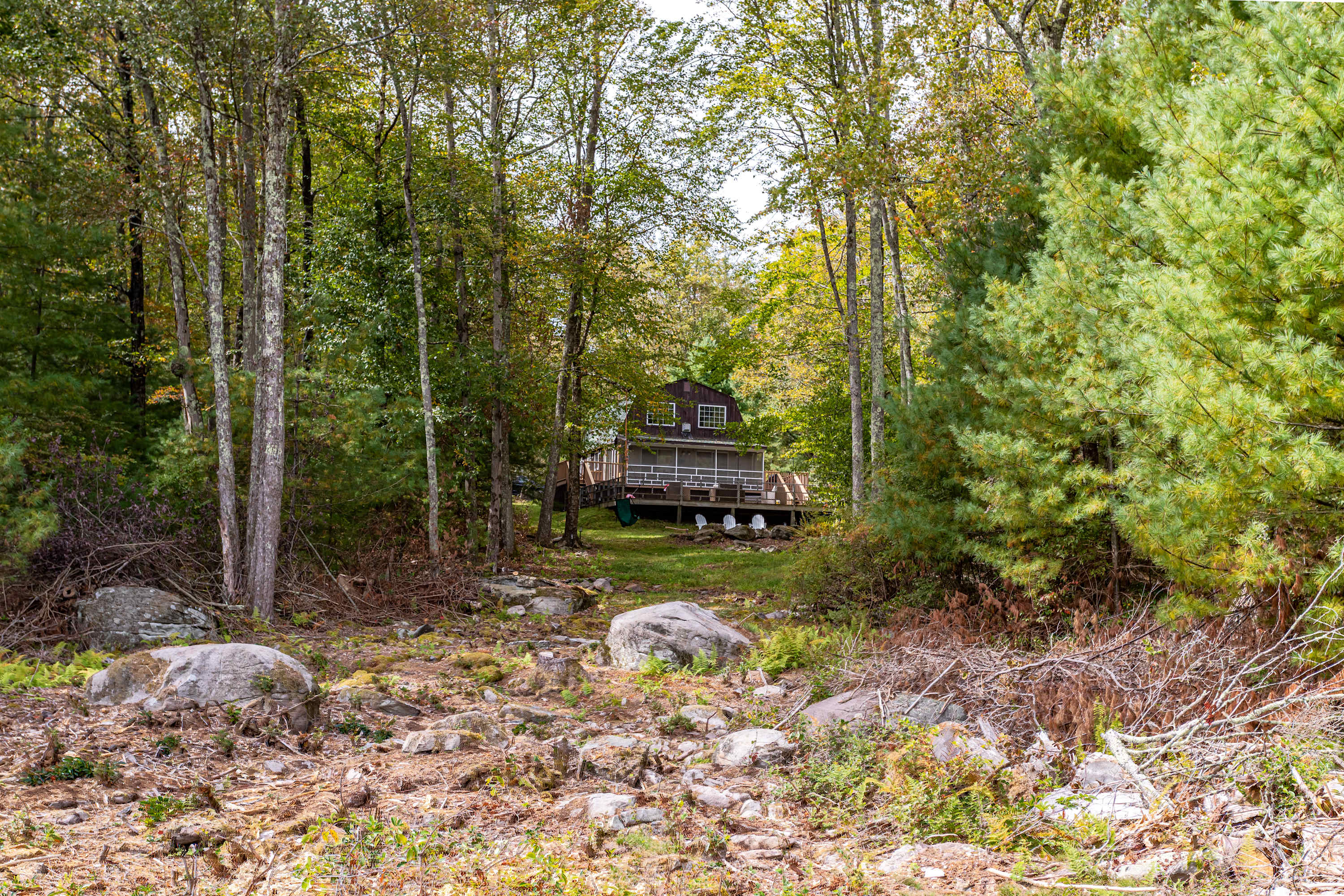 Distant view of the dark brown house, partially obscured by dense green trees and foliage, with a rocky, natural foreground. The house features a screened porch and a wooden deck, blending into the wooded landscape.