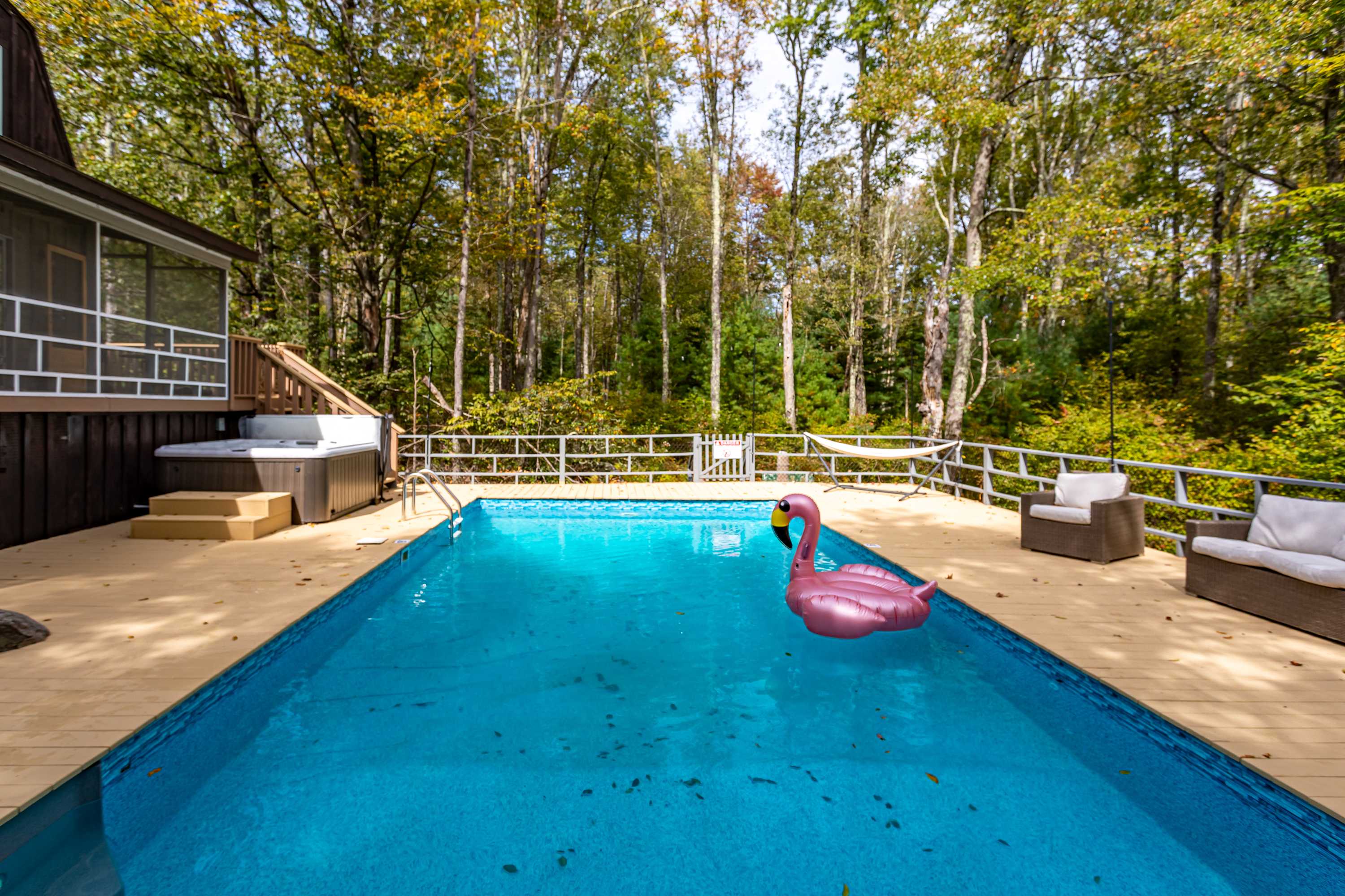 A rectangular swimming pool with a pink flamingo float is surrounded by a light-colored wooden deck, adjacent to a hot tub and the dark brown exterior of the house, all set against a lush green forest.