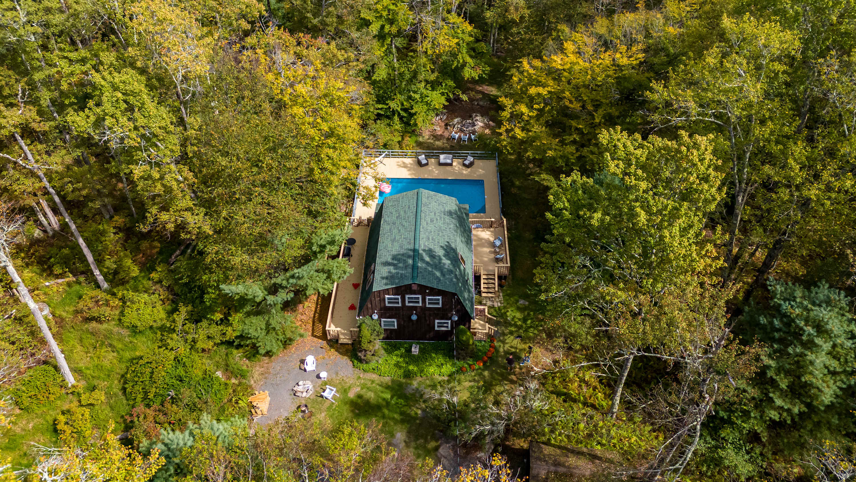 Aerial view of a dark brown barn-style house with a green roof, surrounded by lush green and yellow autumn trees. A large rectangular swimming pool with a light-colored wooden deck is visible next to the house.