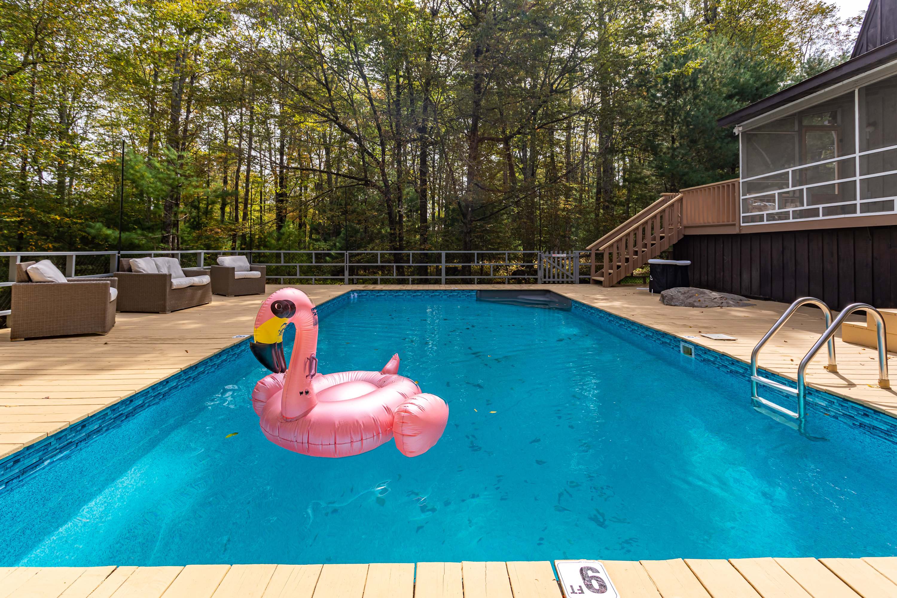 A clear blue swimming pool with a pink flamingo float takes center stage, surrounded by a light-colored wooden deck, with the dark brown house and its screened porch visible in the background through the trees.