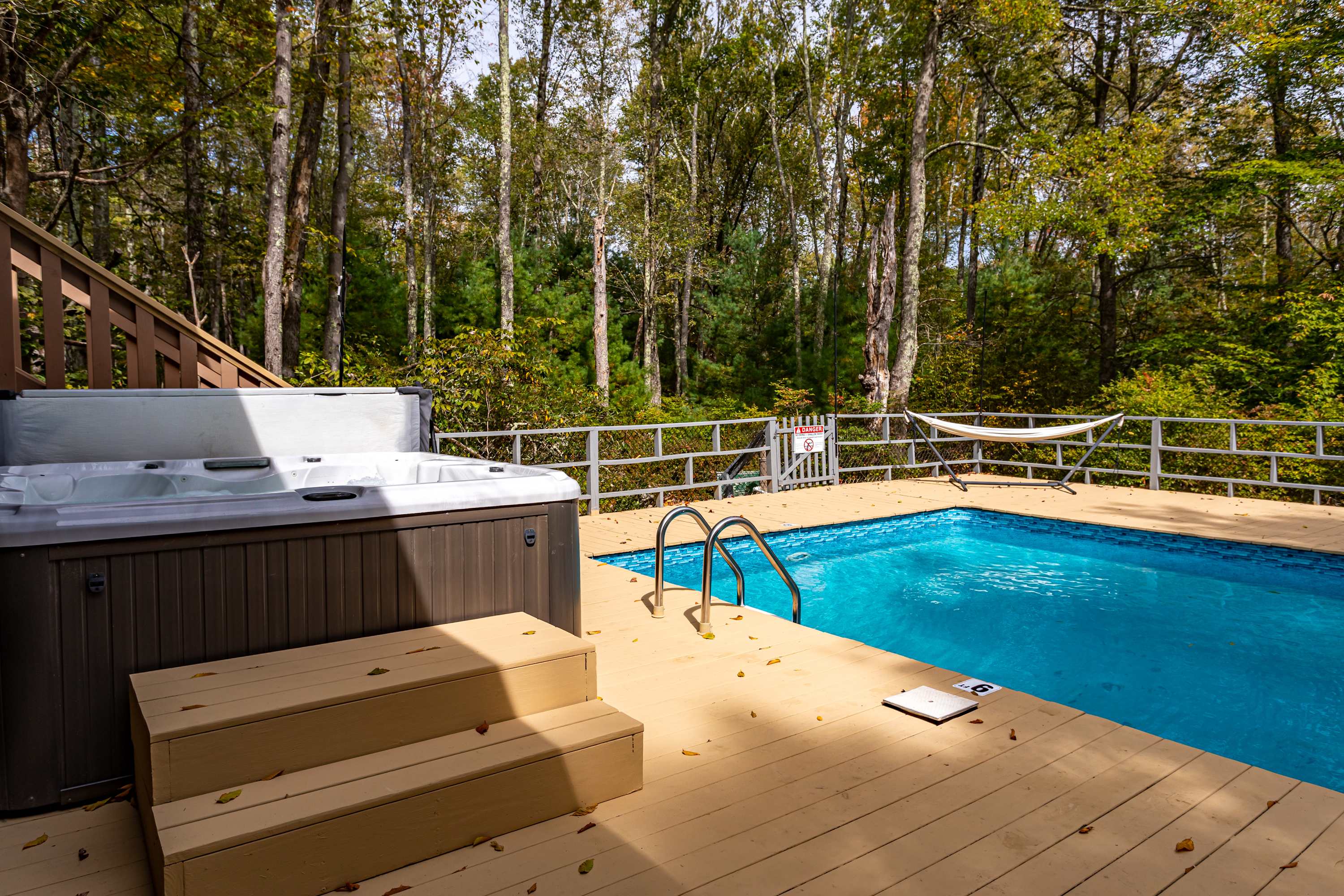 Close-up view of the swimming pool steps and a hot tub on a light-colored wooden deck, surrounded by a metal fence and lush forest. The hot tub has wooden steps leading up to it.