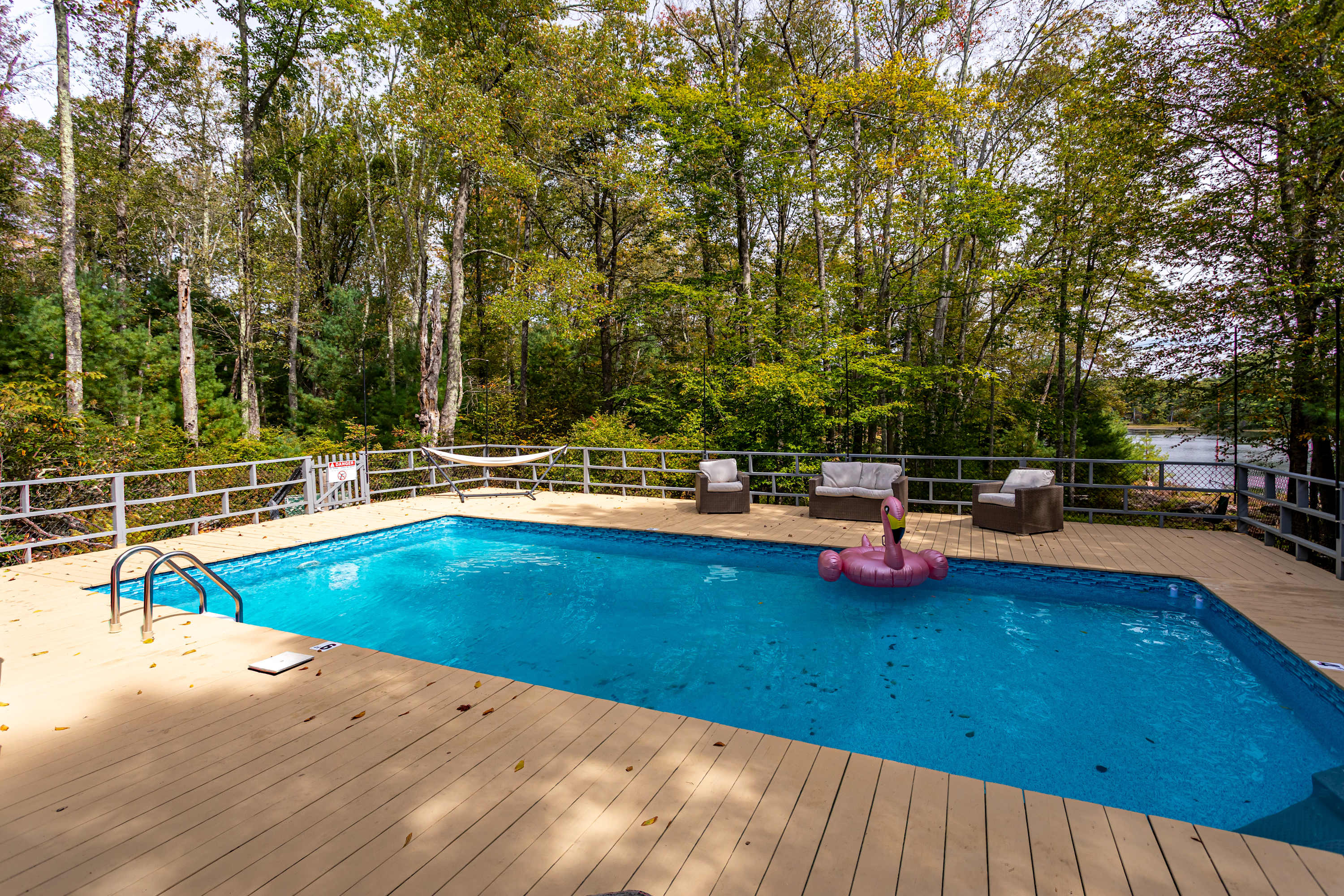 Another wide shot of the rectangular swimming pool with a pink flamingo float, surrounded by a light-colored wooden deck and a metal fence. Outdoor lounge seating and a hammock are visible, set against a backdrop of tall trees.