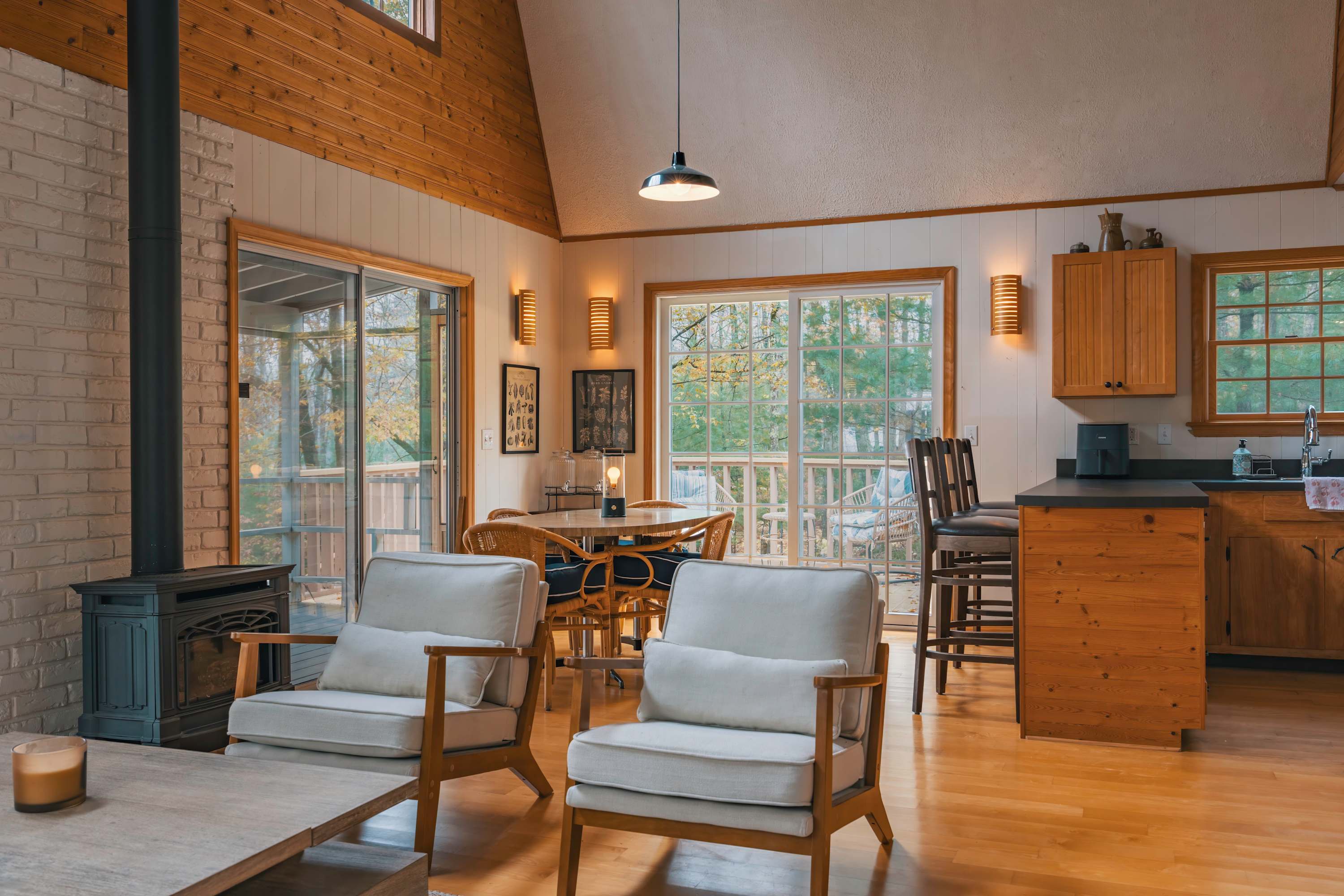 Open-concept living and dining area with light wood floors and white paneled walls, featuring a black wood-burning stove. Sliding glass doors lead to an outdoor deck, while a round dining table and two comfortable armchairs furnish the space.