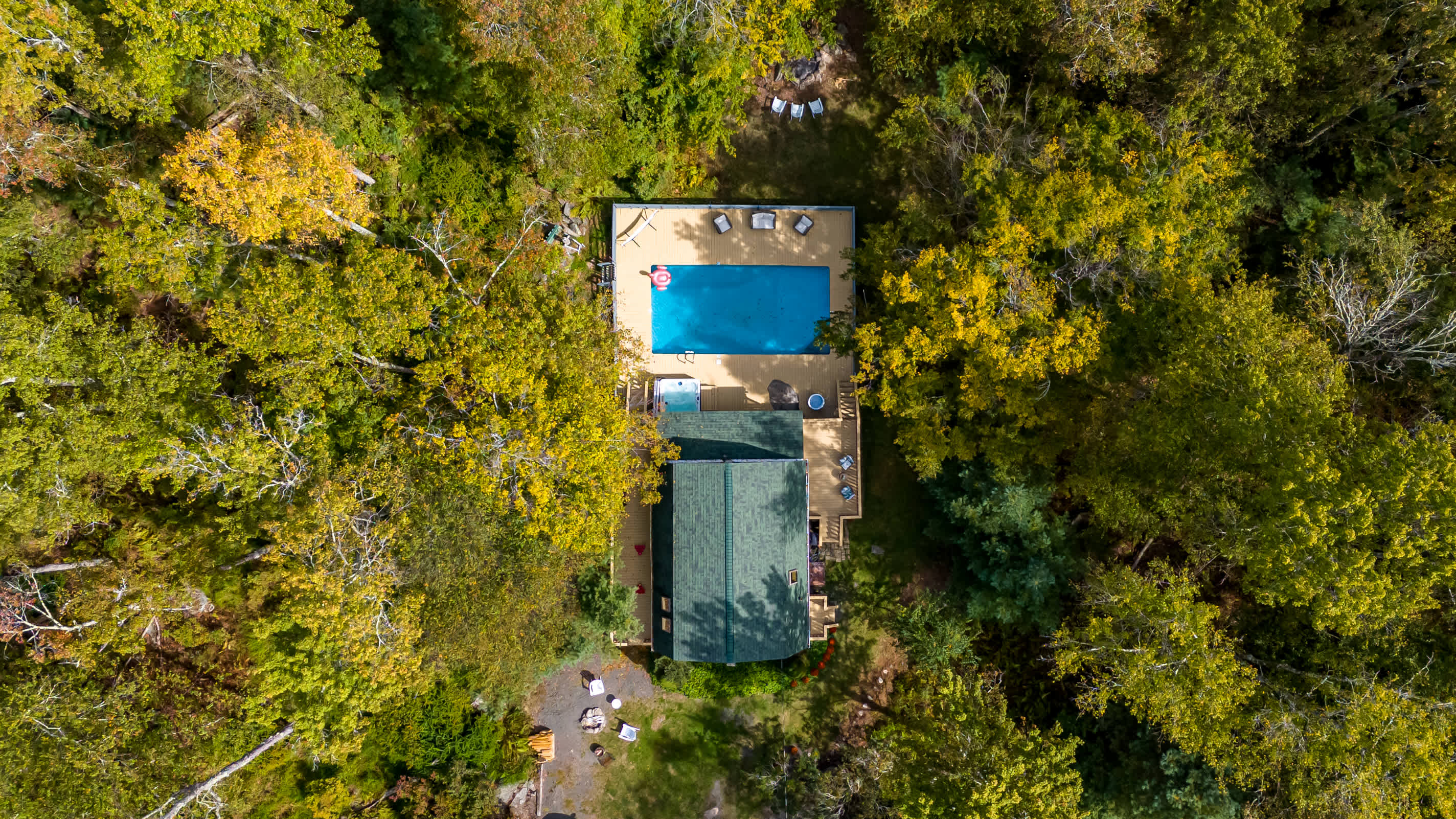An aerial view showcases a rectangular house with a dark roof, connected to a large wooden deck featuring a bright blue swimming pool with a pink float and a hot tub, all nestled within a dense, vibrant green and yellow forest.