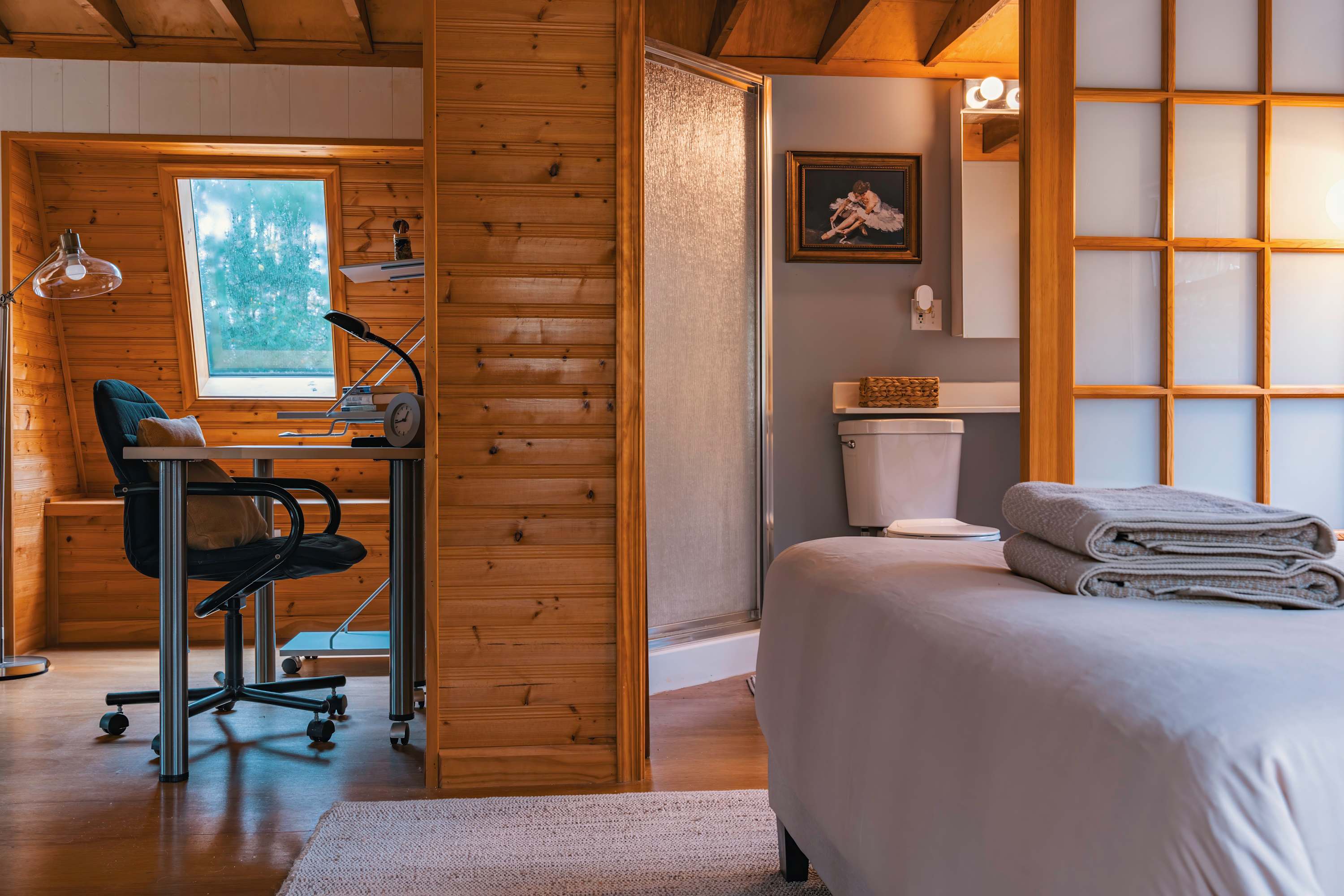 Bright bedroom with a queen bed on the right, an office nook on the left with a desk and ergonomic chair, all set against warm wood paneling and exposed ceiling beams, with a glimpse of a modern bathroom.