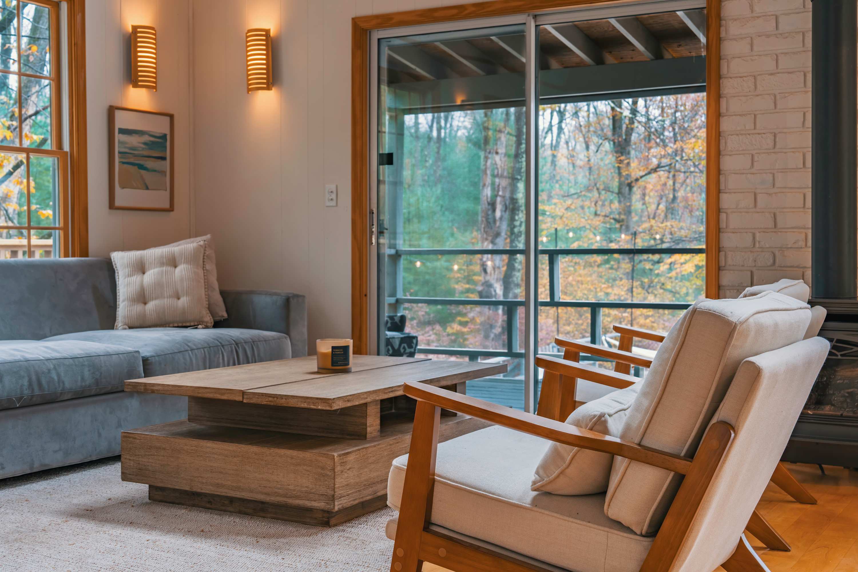 Inviting living room with a grey sofa, two light-colored armchairs, and a rustic wooden coffee table, featuring large sliding glass doors that open to a deck with views of autumn trees.