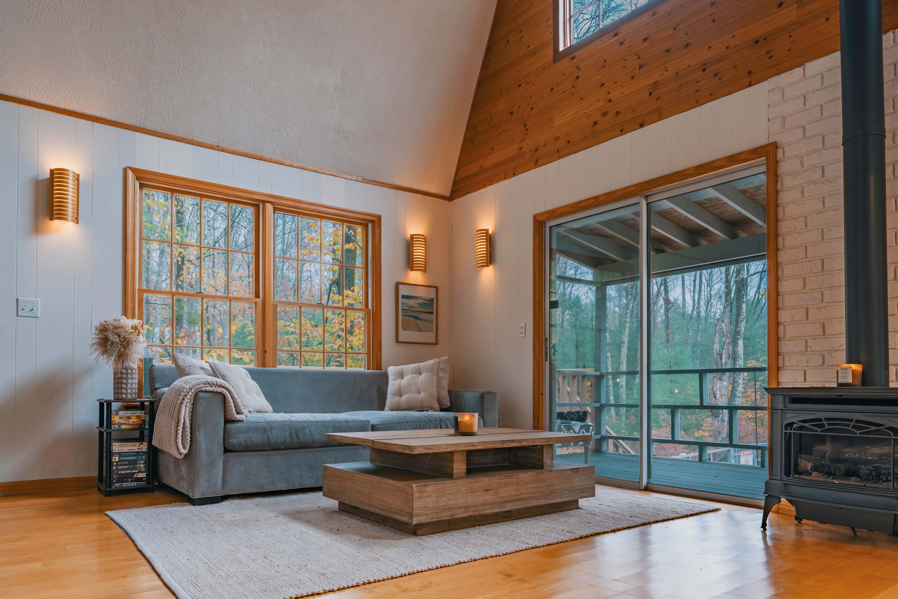 Inviting living room with a comfortable grey sofa, a rustic wooden coffee table, a wood-burning stove, and large windows offering views of the surrounding autumn trees, creating a cozy ambiance.