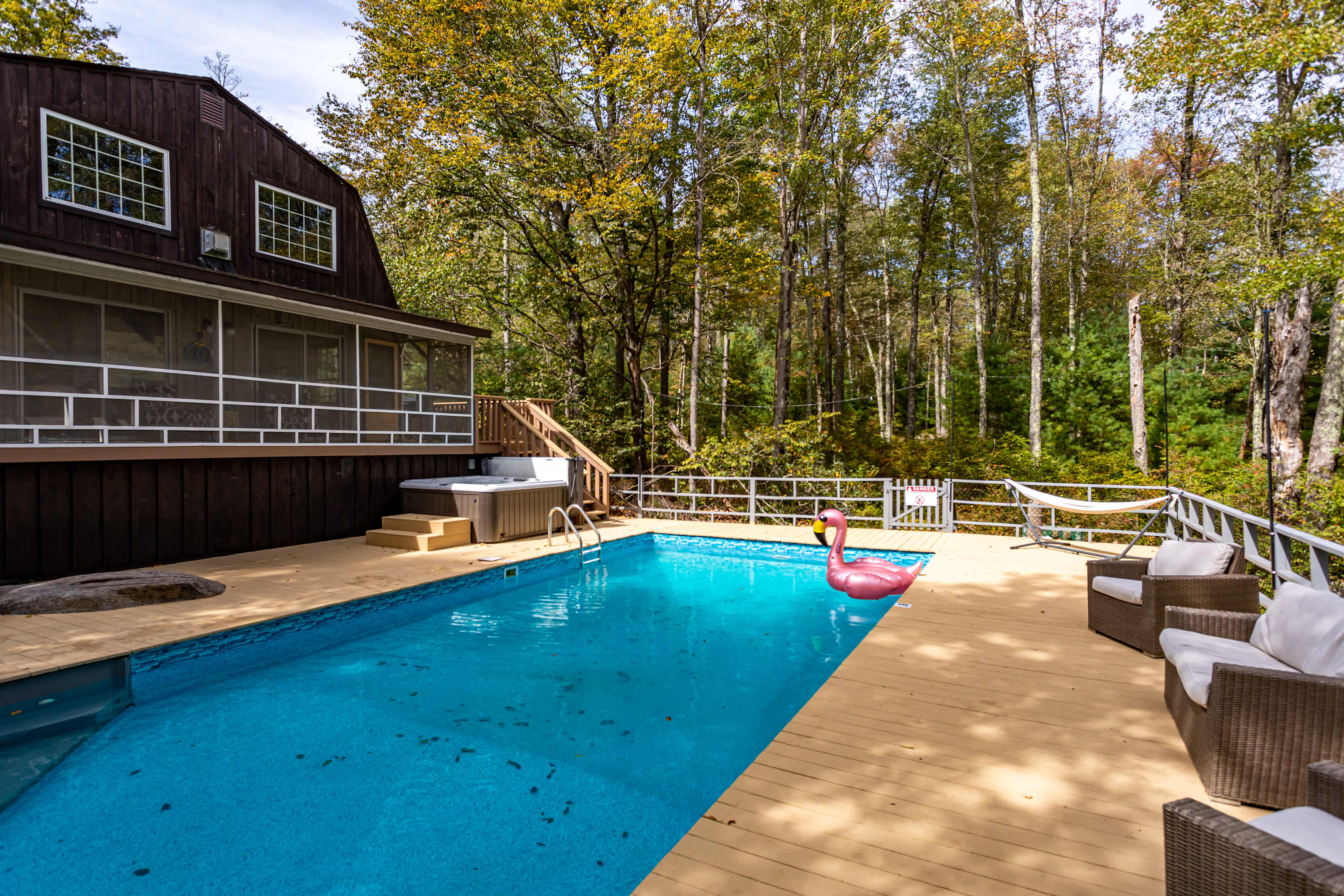 A vibrant blue swimming pool with a pink flamingo float is centered on a spacious light-colored wooden deck, flanked by a hot tub and the dark brown house with a screened porch, all nestled within a dense green forest.