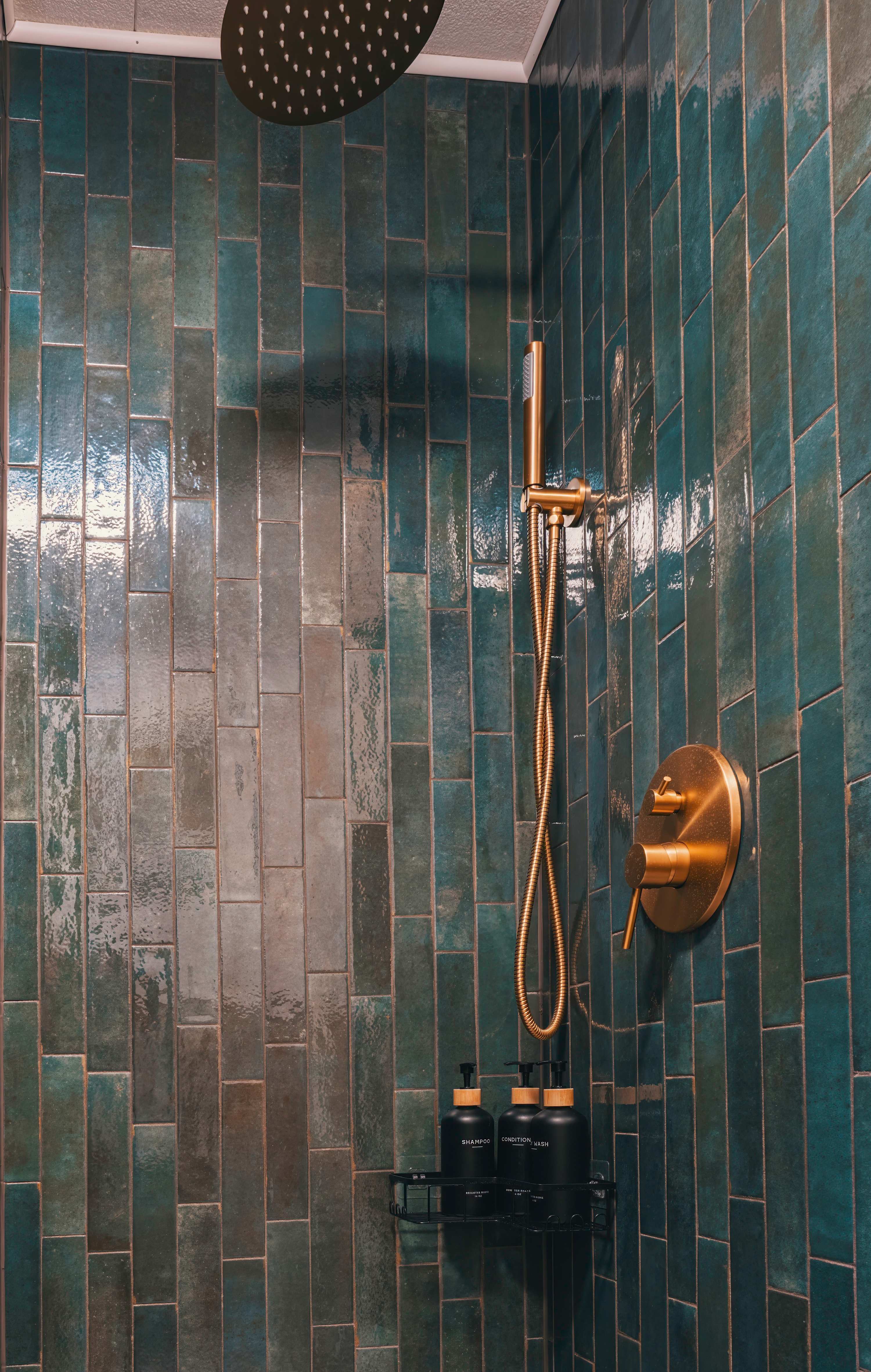Close-up of a stylish shower with dark teal subway tiles, featuring a gold rain shower head and a matching handheld shower, with black bottles of shampoo and conditioner on a shelf.