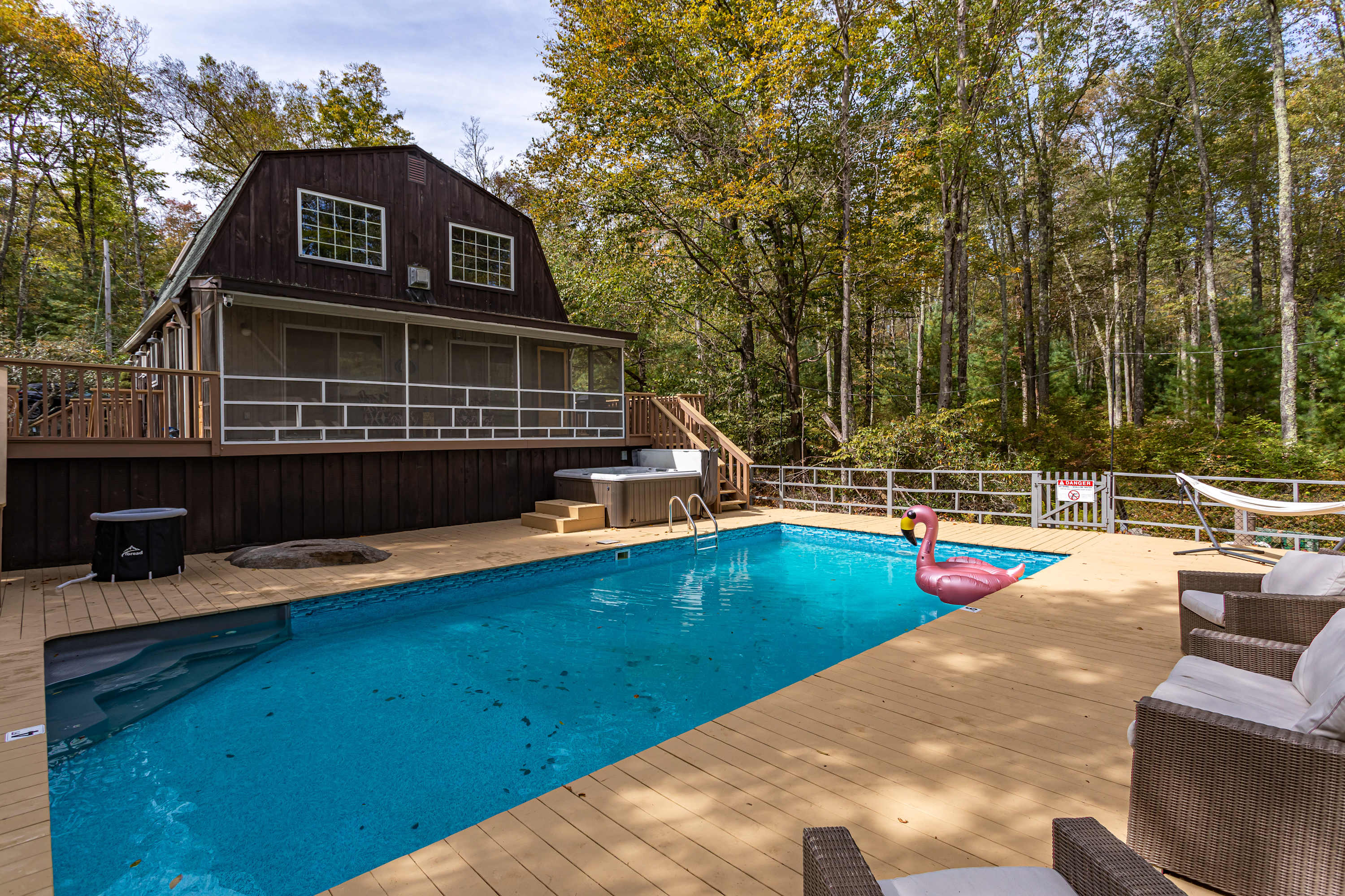 A bright blue swimming pool with a pink flamingo float anchors a large light-colored wooden deck, featuring a hot tub and outdoor seating, next to the dark brown house with a screened porch, surrounded by a verdant forest.